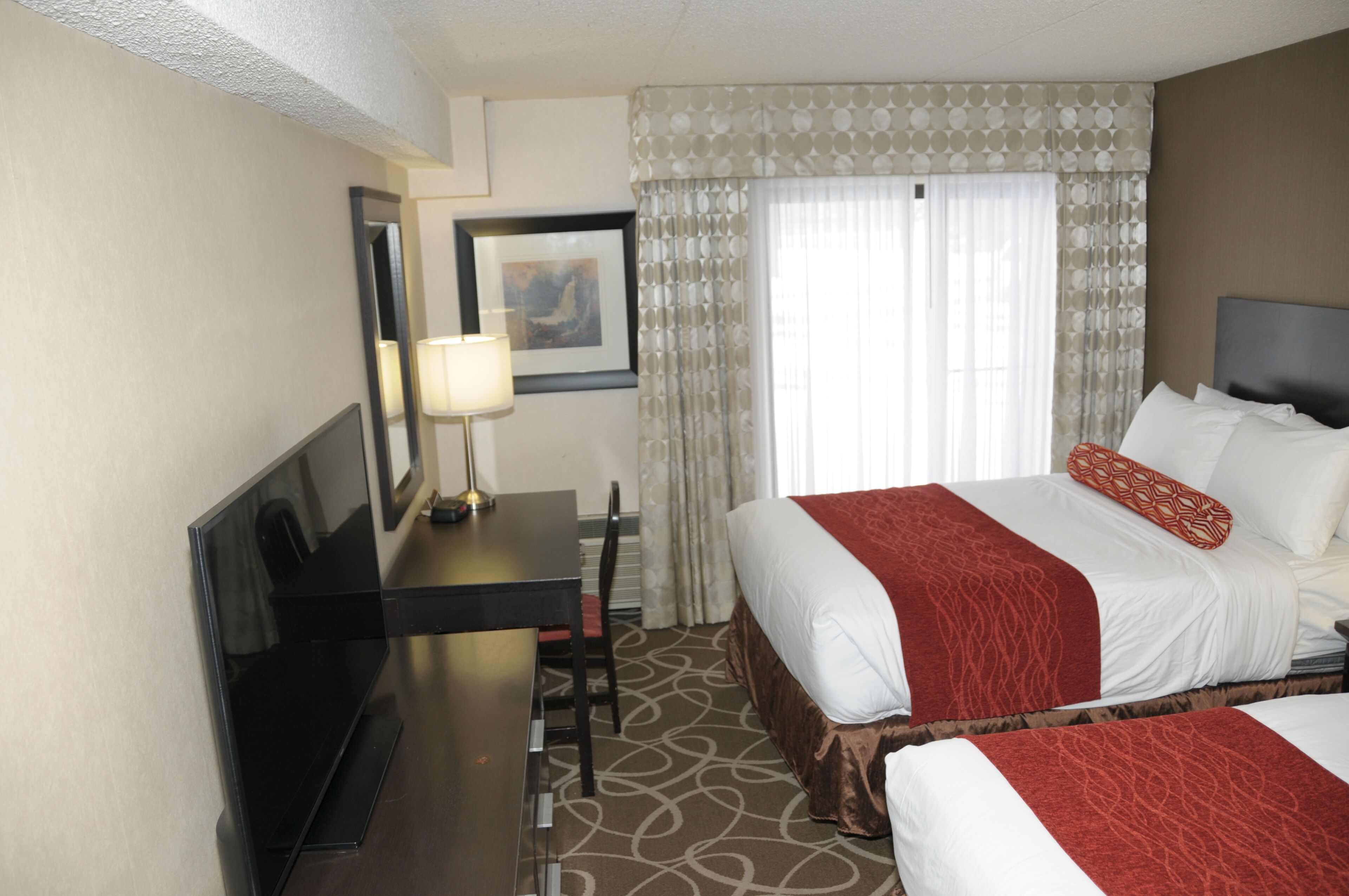 Una habitación de hotel con cama, escritorio, silla, televisor y ventana con cortinas.