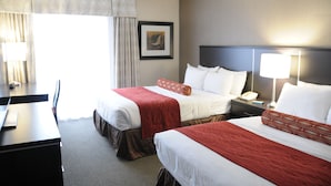 A hotel room with two beds, a desk, a chair, a nightstand with a lamp, and a framed picture on the wall.