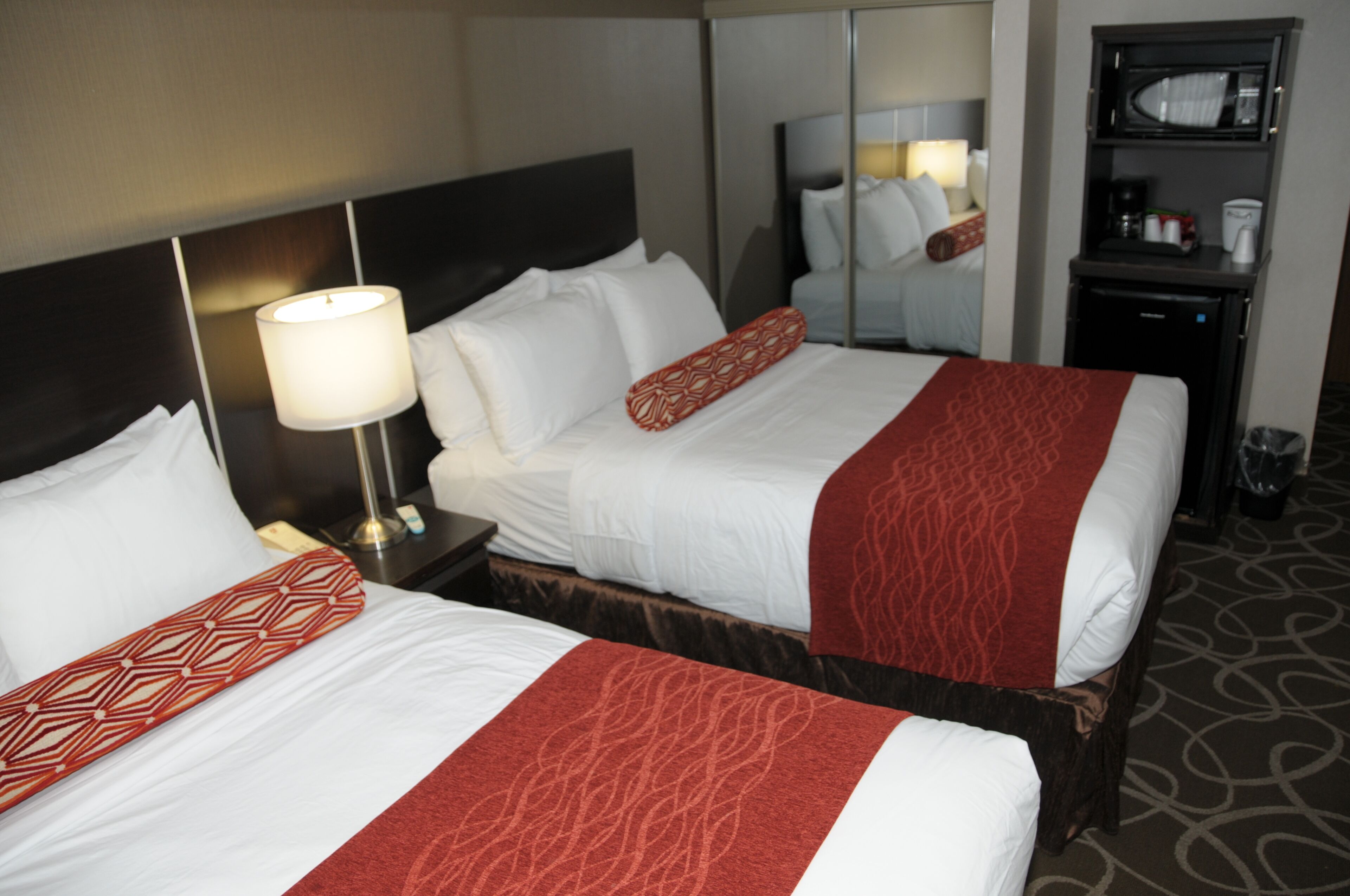 Een hotelkamer met twee bedden, elk voorzien van witte lakens en rode sierkussens. Er staat een nachtkastje met een lamp en een klein tafeltje met een koffiezetapparaat en kopjes. Boven de bedden hangt een spiegel aan de muur.