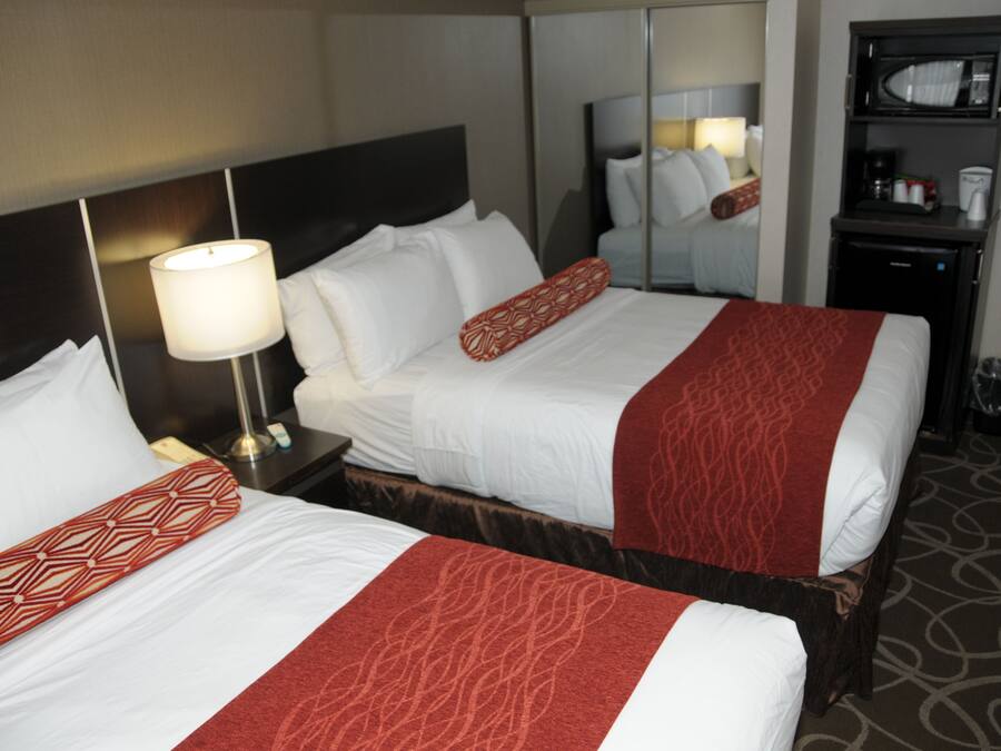 Een hotelkamer met twee bedden, elk voorzien van witte lakens en rode sierkussens. Er staat een nachtkastje met een lamp en een klein tafeltje met een koffiezetapparaat en kopjes. Boven de bedden hangt een spiegel aan de muur.