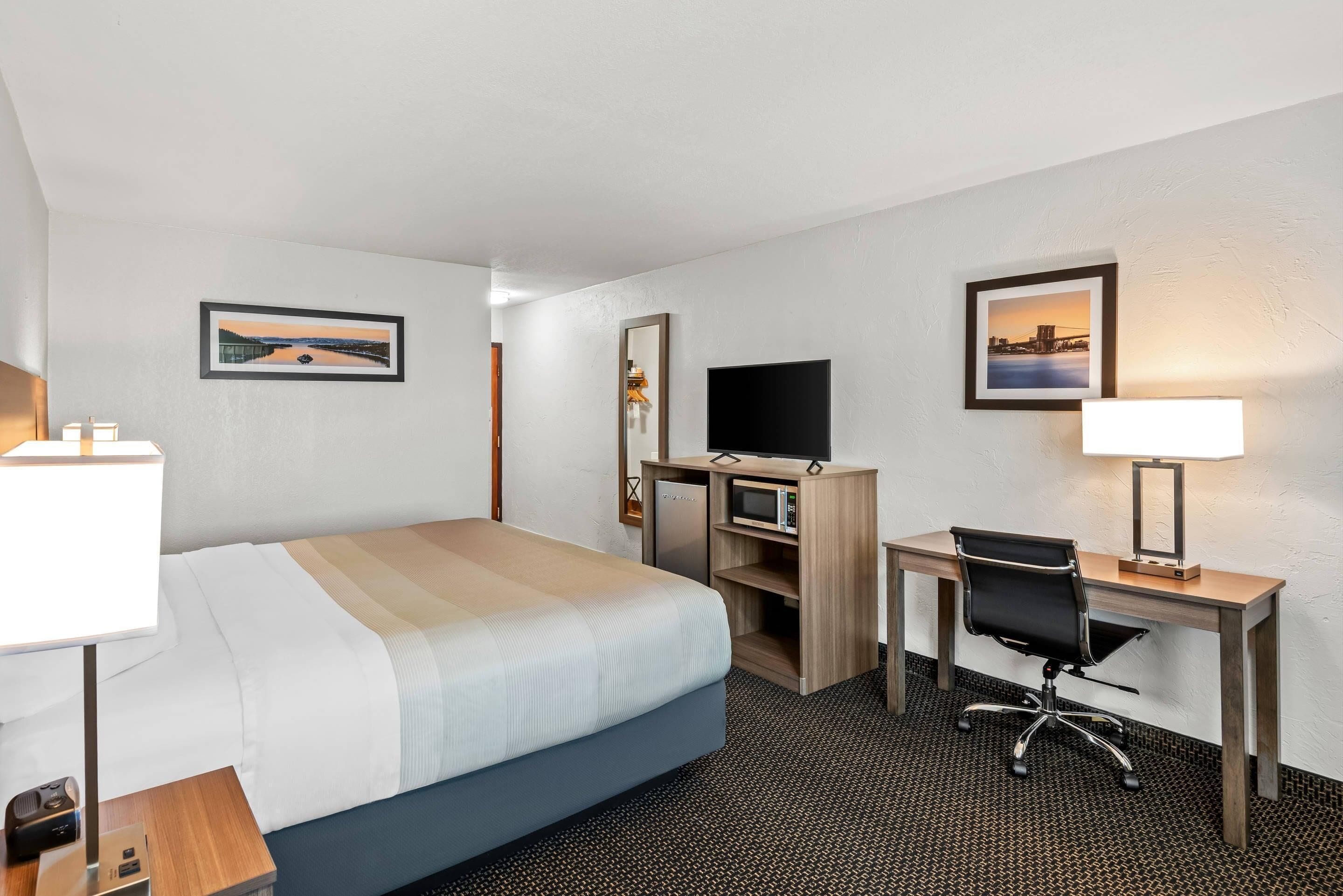 Una habitación de hotel con cama, escritorio, silla, televisor y dos lámparas fijadas a la pared.