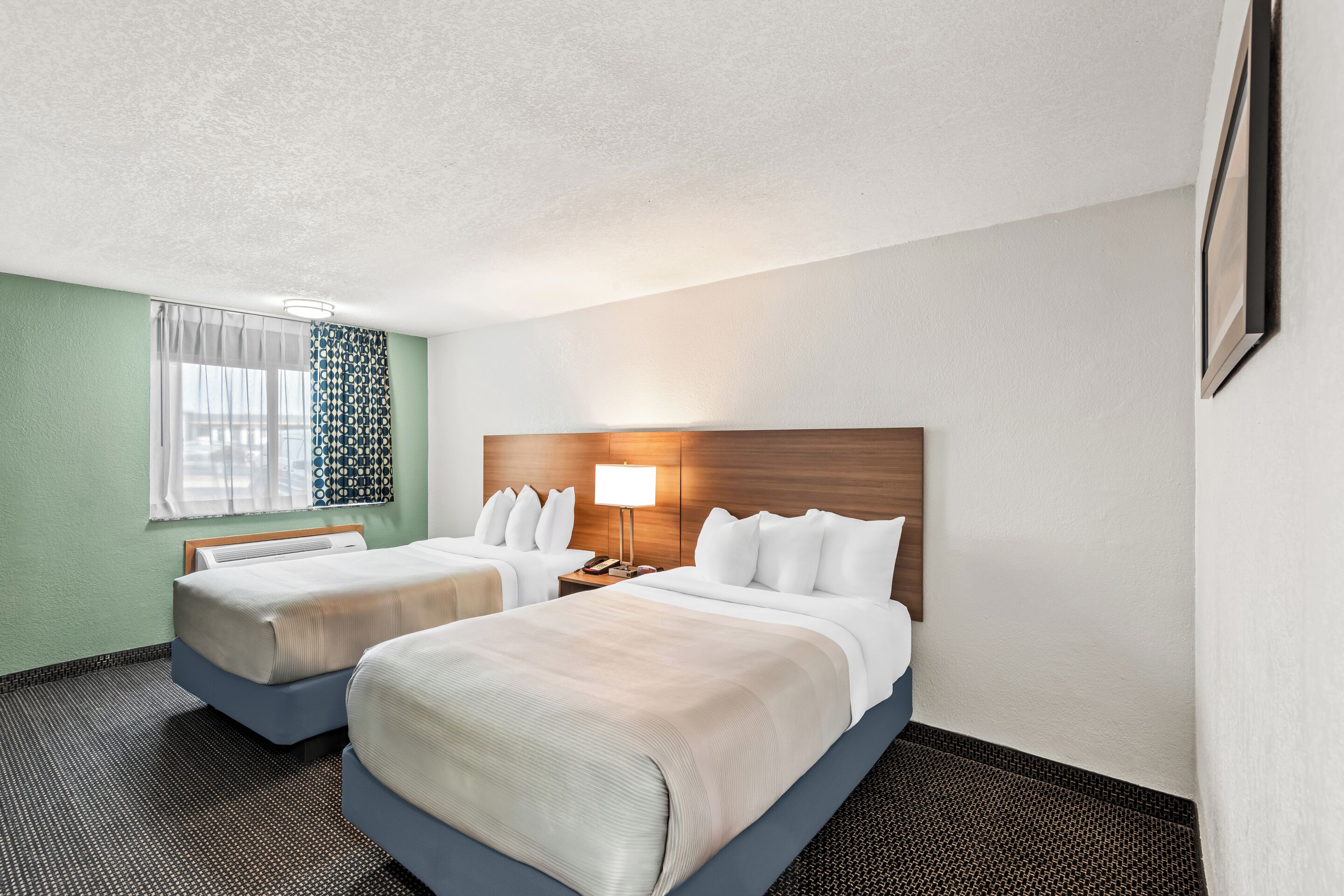 A hotel room with two beds, a wooden headboard, a nightstand with a lamp, a window with patterned curtains, and a framed picture on the wall.
