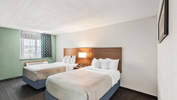 A hotel room with two beds, a wooden headboard, a nightstand with a lamp, a window with patterned curtains, and a framed picture on the wall.