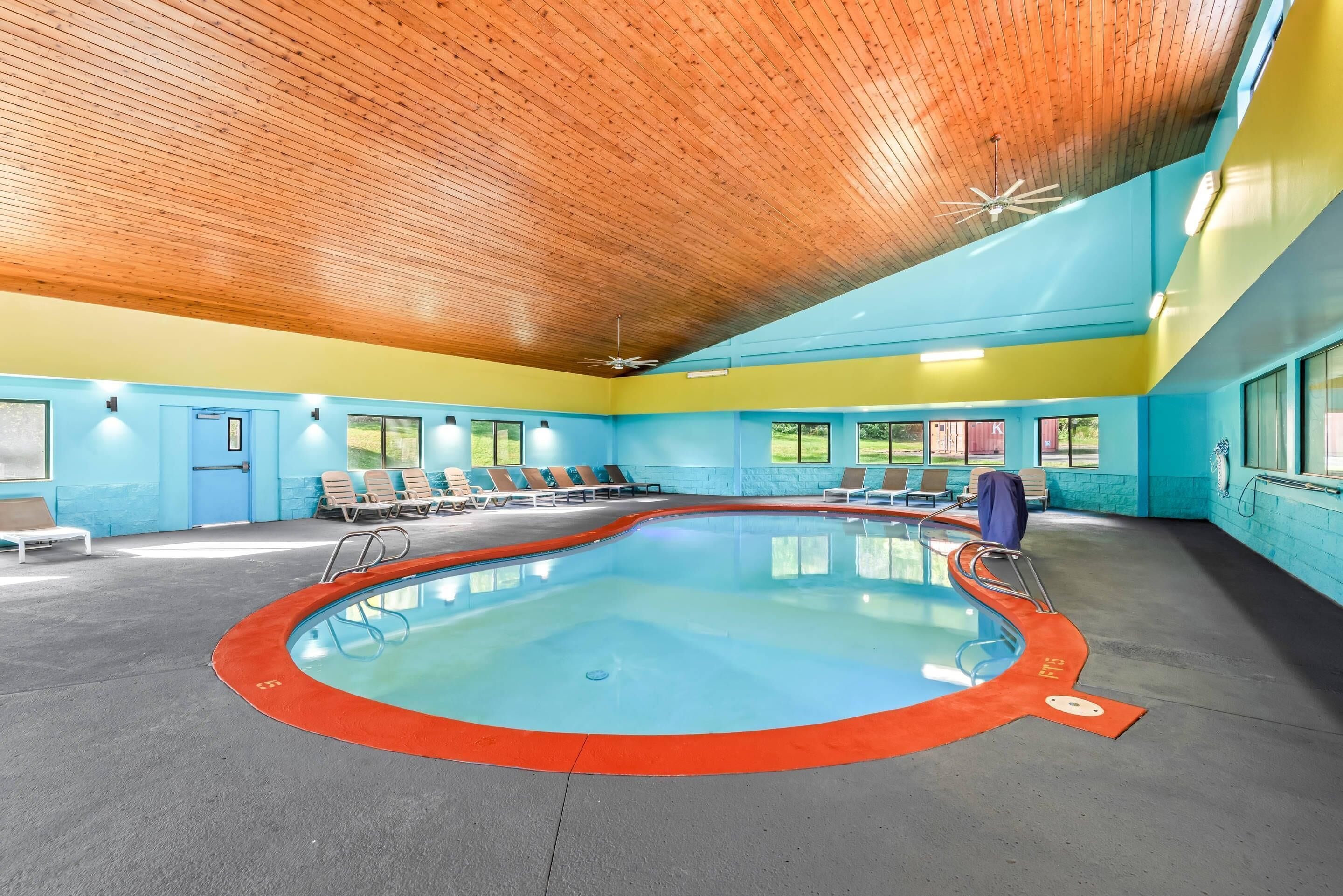 Una piscina interior con techo de madera, paredes azules y ventanas iluminadas por el sol.