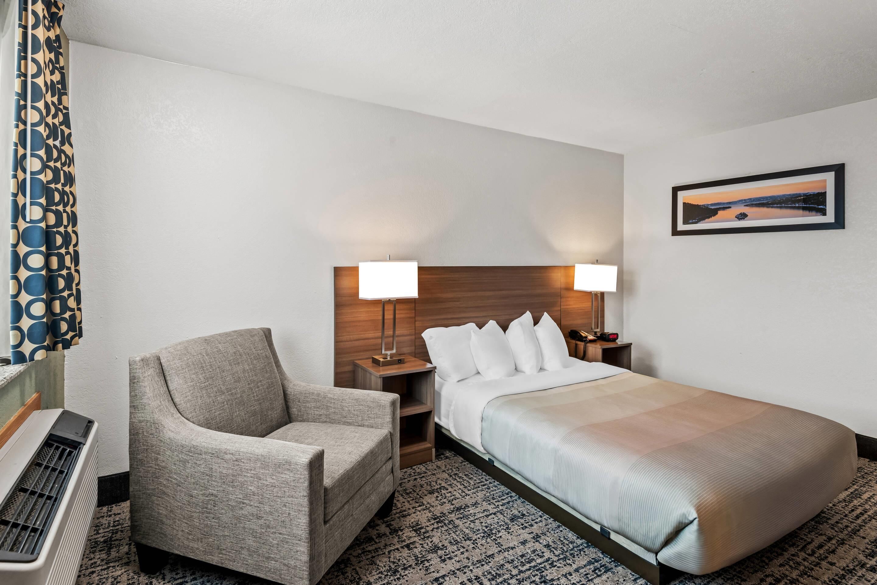 A hotel room with a bed, two bedside lamps, a chair, a small table, and a wall-mounted picture.