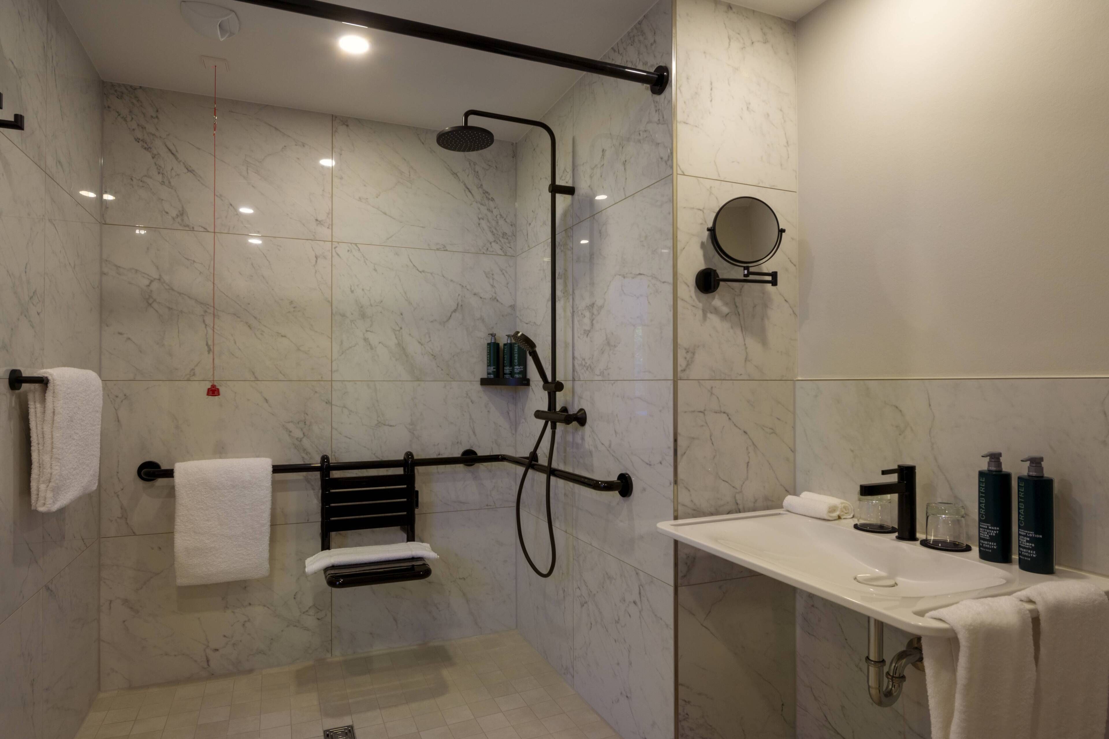 Une salle de bain moderne avec une douche à parois de verre, un porte-serviettes noir, un lavabo blanc avec des articles de toilette et un miroir rond.