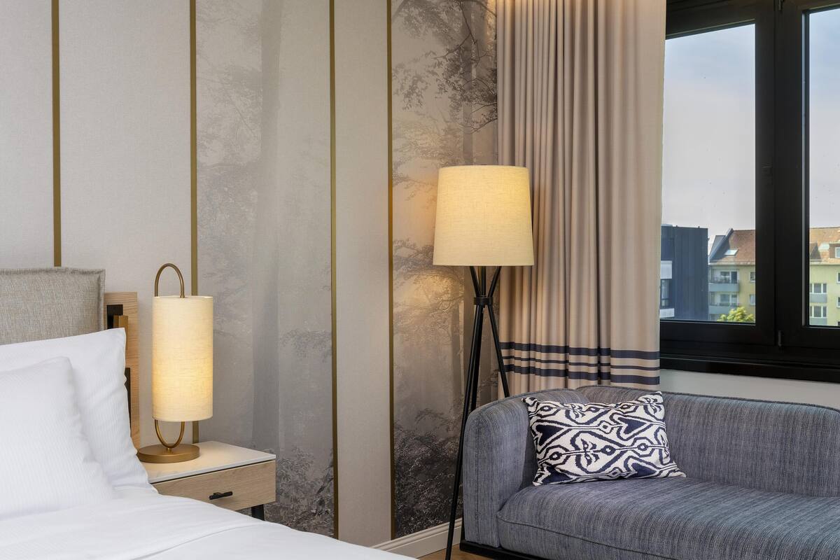 Habitación de hotel con cama, mesita de noche con lámpara, sofá con cojines estampados y ventana con vistas a edificios.