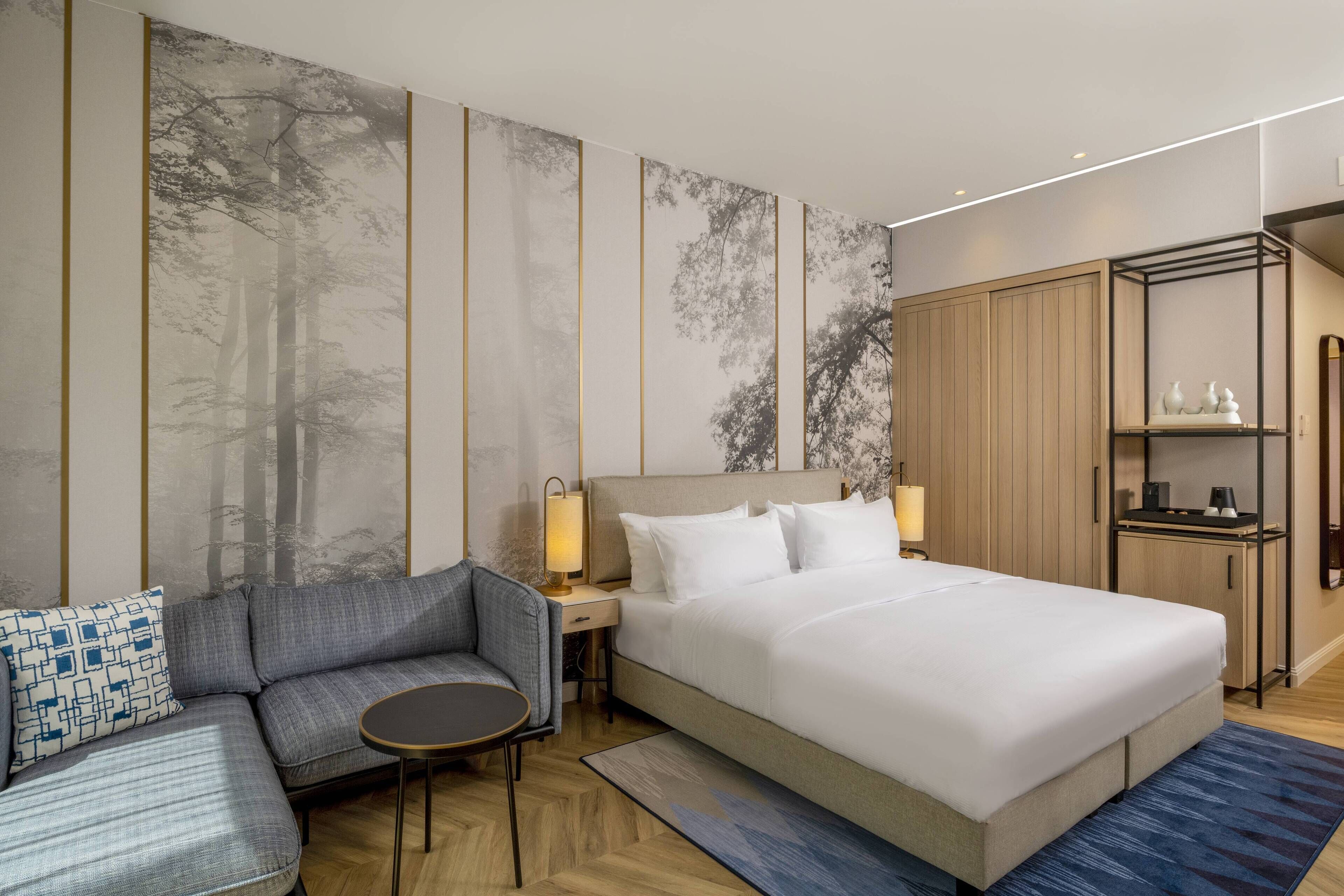 A modern hotel room with a large bed, a sofa, a small table, and a wardrobe.