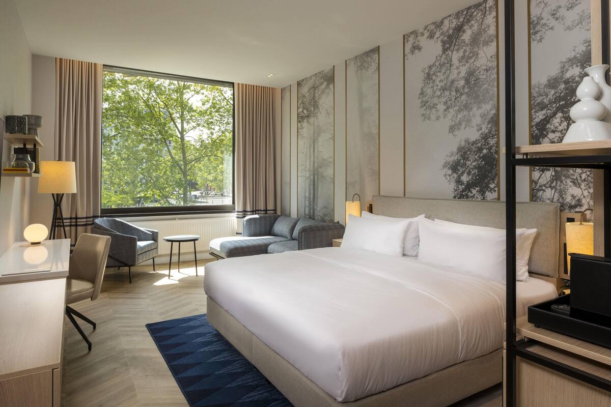 Habitación de hotel moderna con una cama grande, un escritorio, una silla y vistas a árboles por la ventana.
