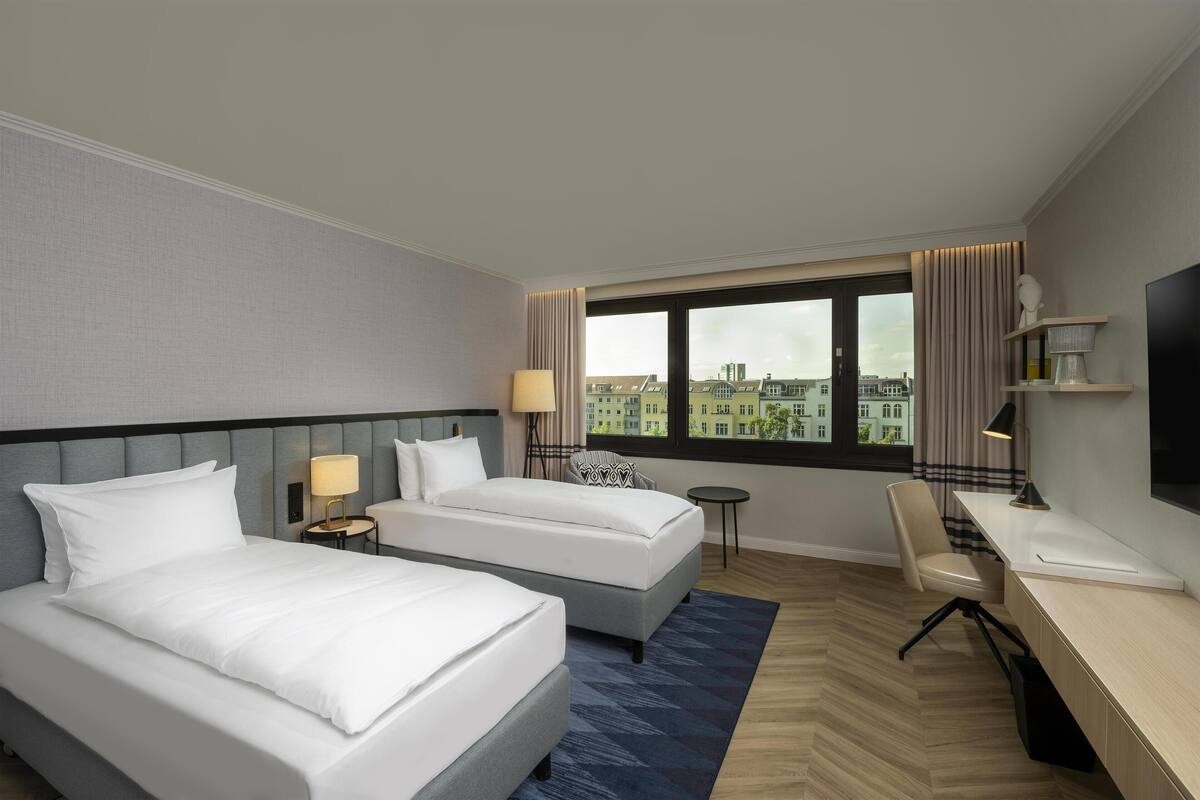 Habitación de hotel con dos camas, un escritorio, una silla, un televisor y vistas a edificios a través de la ventana.