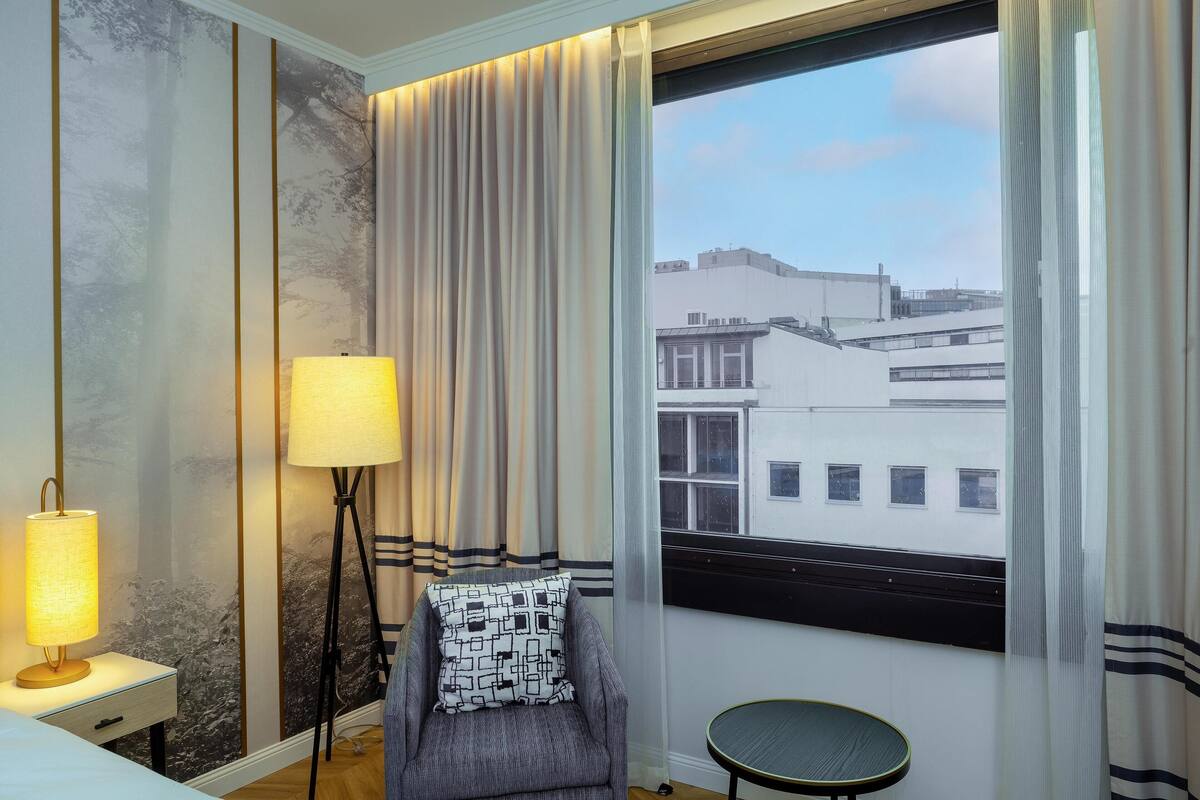Habitación de hotel con vista a edificios, una silla con un cojín estampado, una lámpara de pie y una mesita.
