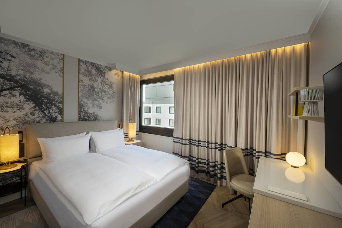 Habitación de hotel con una cama grande, un escritorio, una silla, un televisor y una ventana con cortinas.