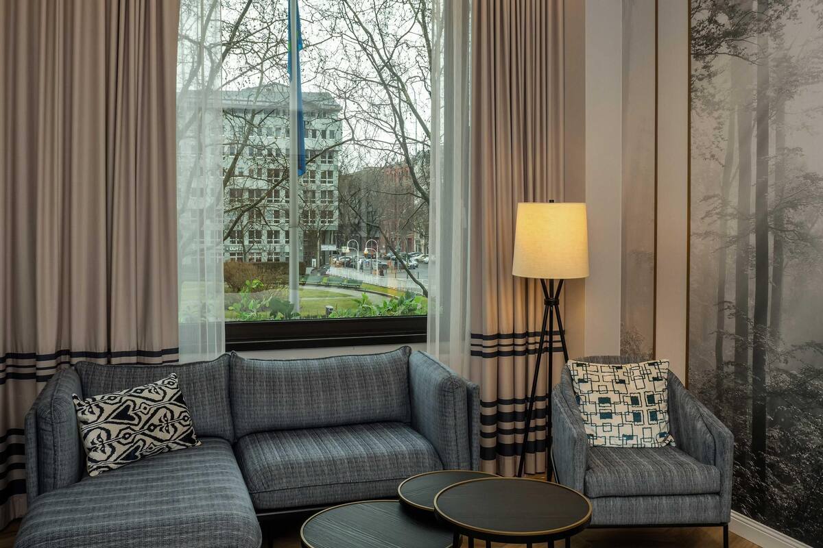 Habitación de hotel moderna con un sofá gris, una pequeña mesa redonda y una silla con cojines estampados. Un gran ventanal ofrece vistas a edificios y árboles.