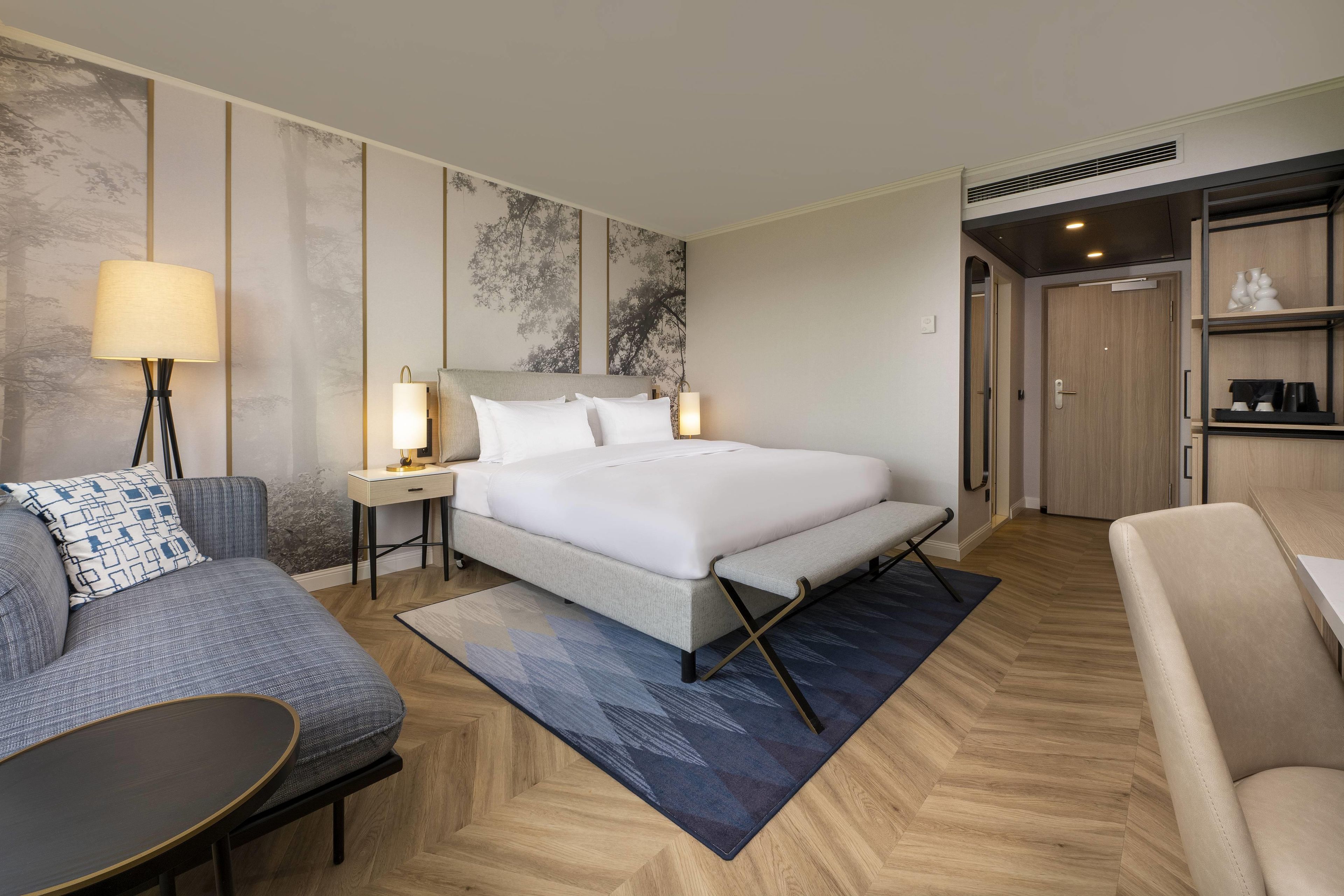 A hotel room with a bed, a desk, a chair, a sofa, and a nightstand with lamps.