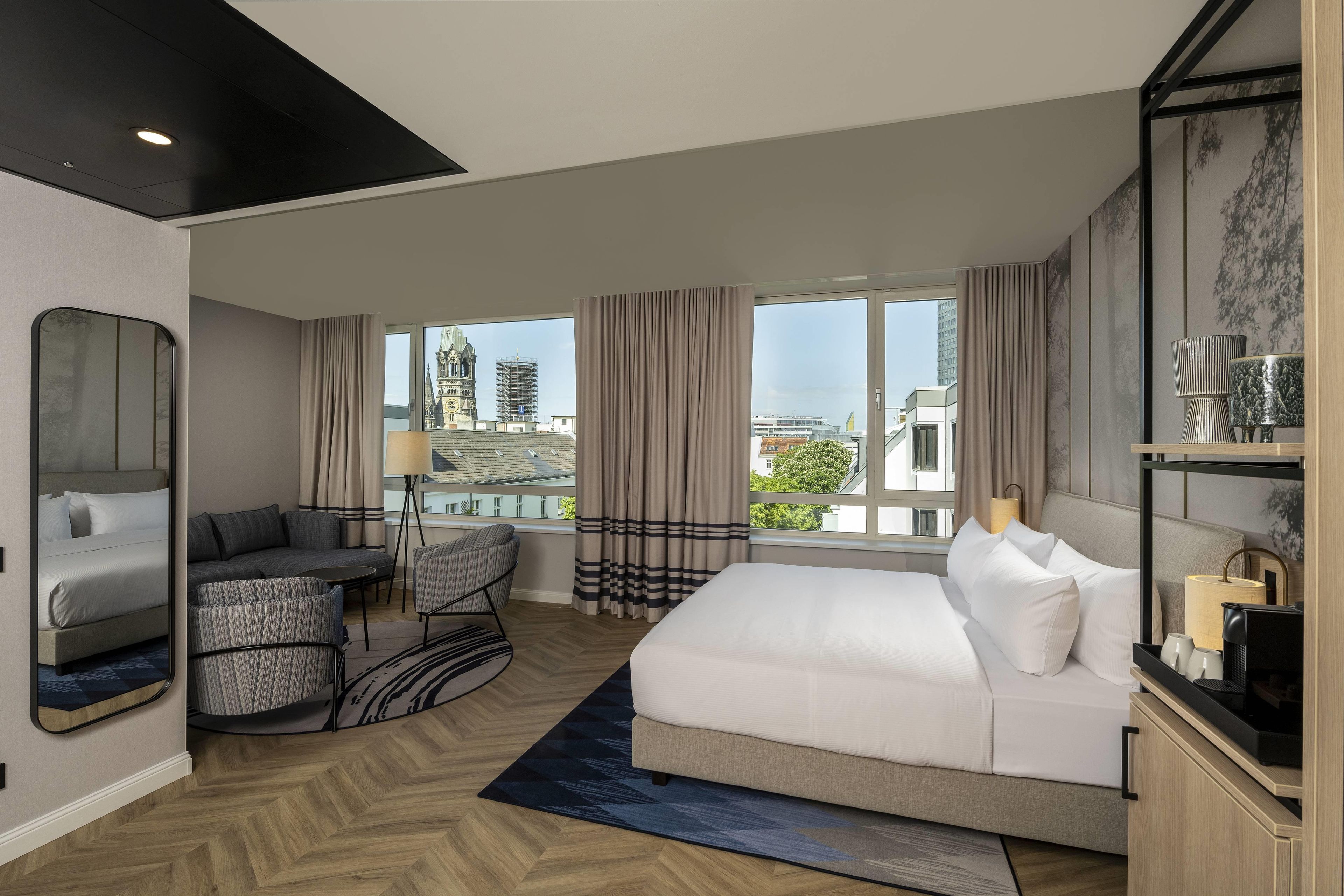 A modern hotel room with a large bed, a seating area with a sofa and armchairs, a small table, and a view of the cityscape through large windows.