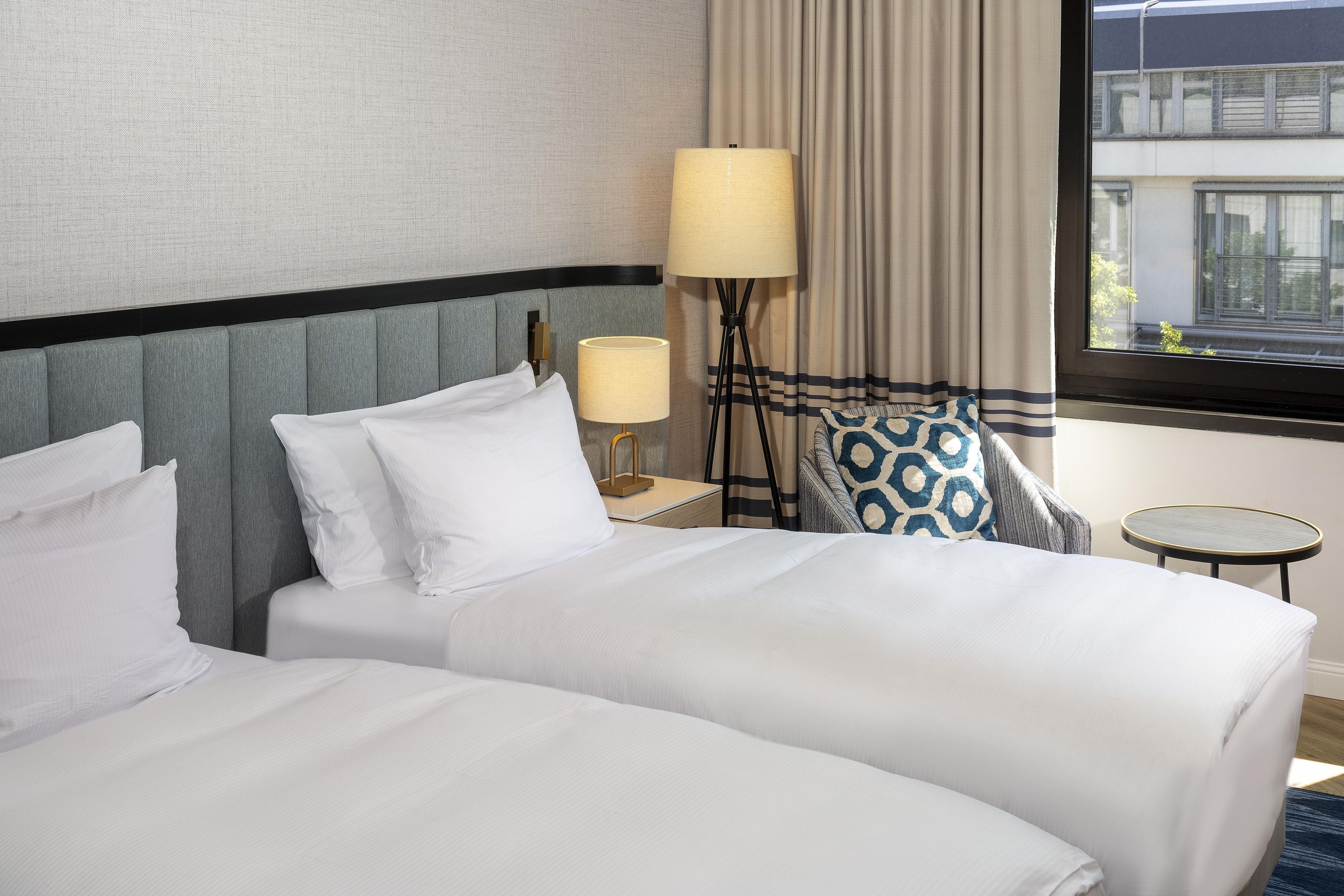 A hotel room with two beds, a lamp, a small table, and a view of the outside.