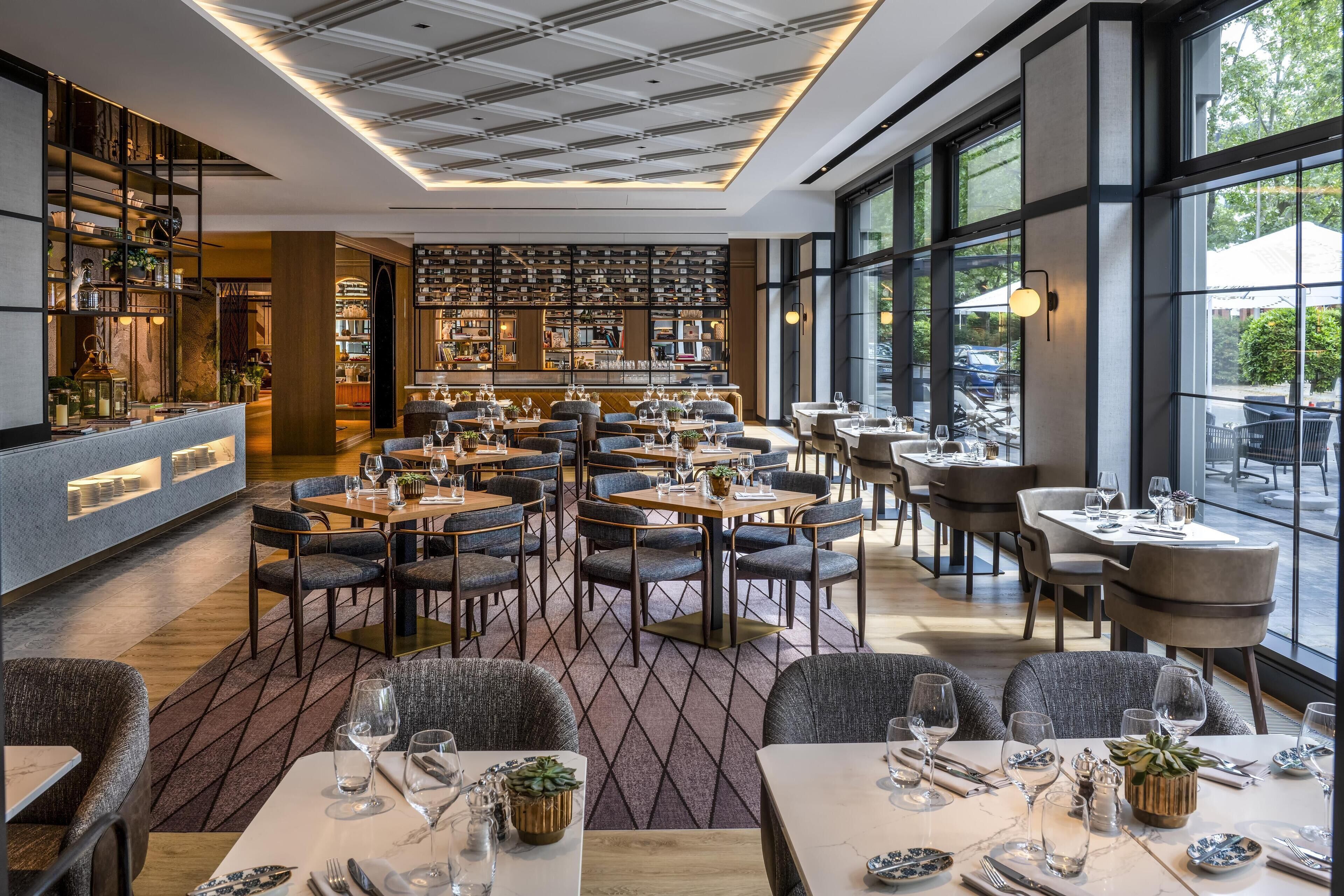 A modern restaurant with multiple dining areas, a bar, and large windows offering views of the outdoors.