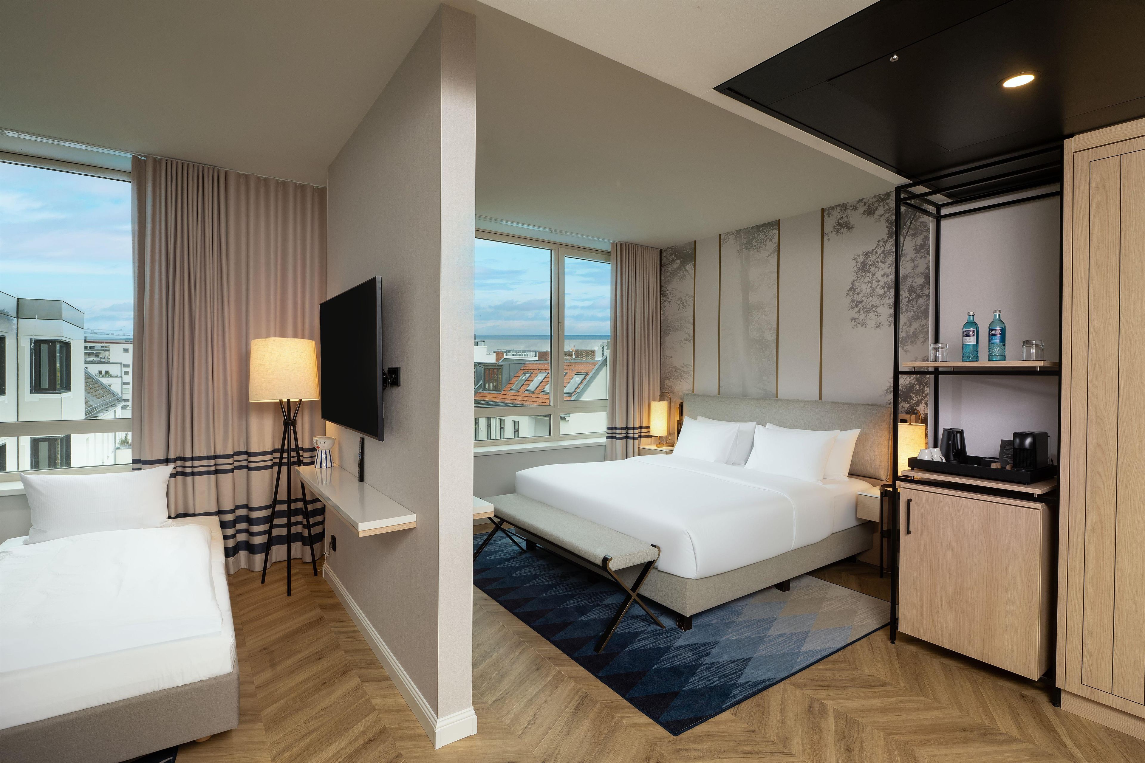 A hotel room with a bed, bedside table, lamp, TV, and a view of the city.