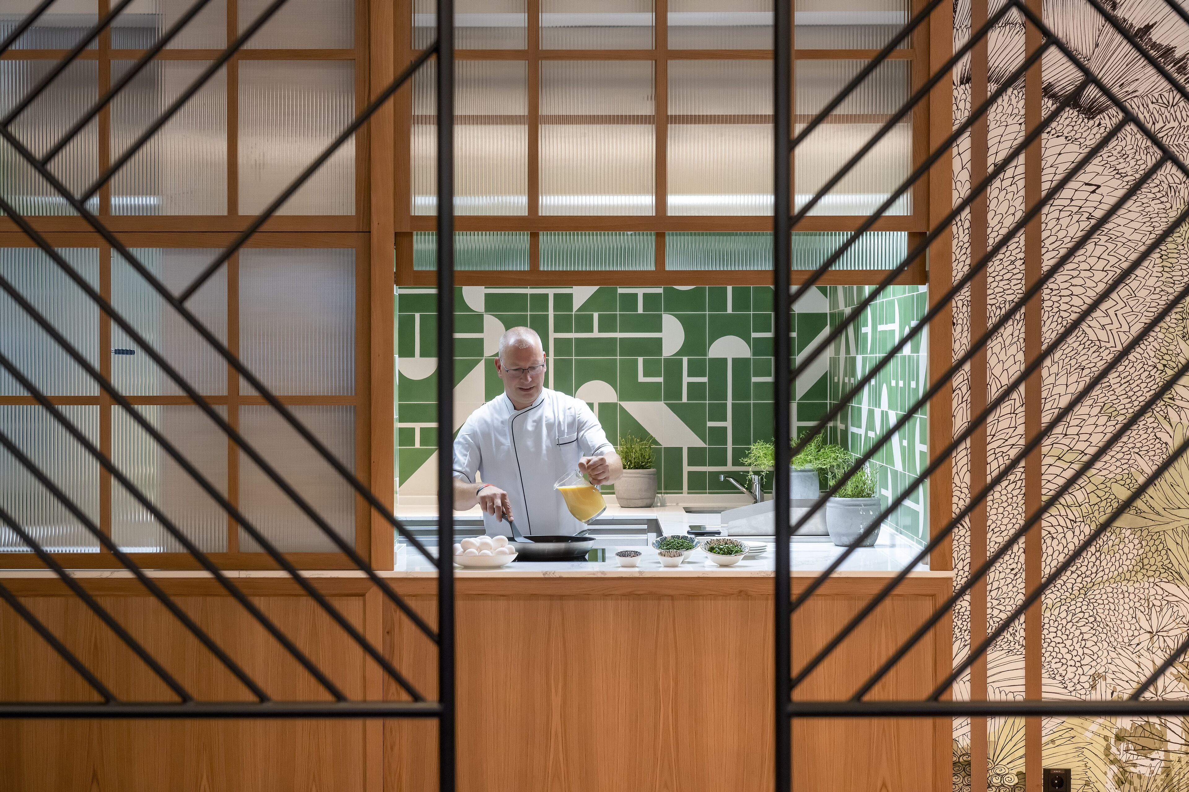 Ein Koch bereitet Essen in einer Küche mit einer grün-weißen Fliesenrückwand und Holzelementen.