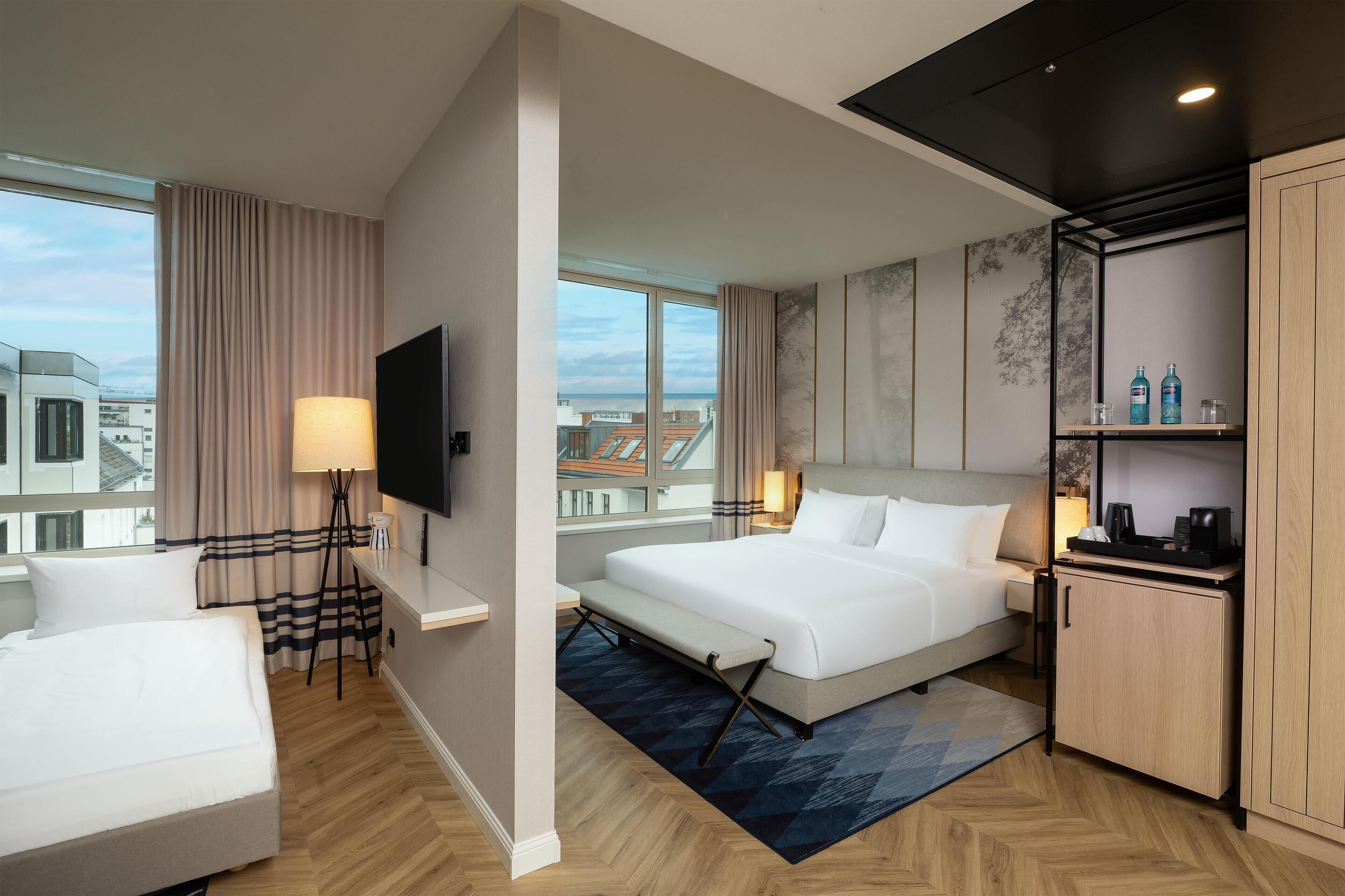 A modern hotel room with a large bed, a desk, a chair, a TV, and a view of the city.