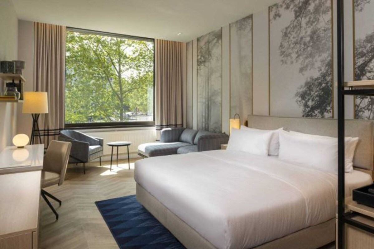 A modern hotel room with a large bed, a desk, a chair, and a sofa. There is a window with a view of trees.