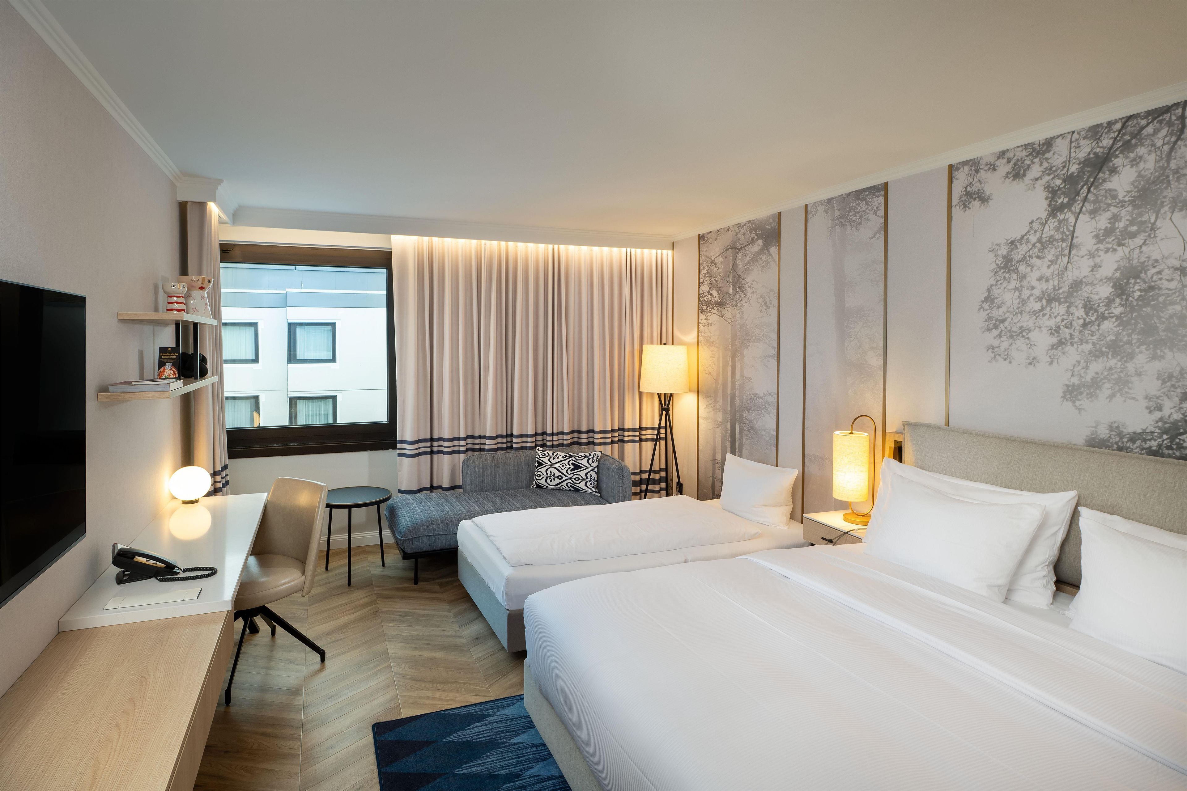 Quarto de hotel com cama, escrivaninha, cadeira, televisão e janela com cortinas.