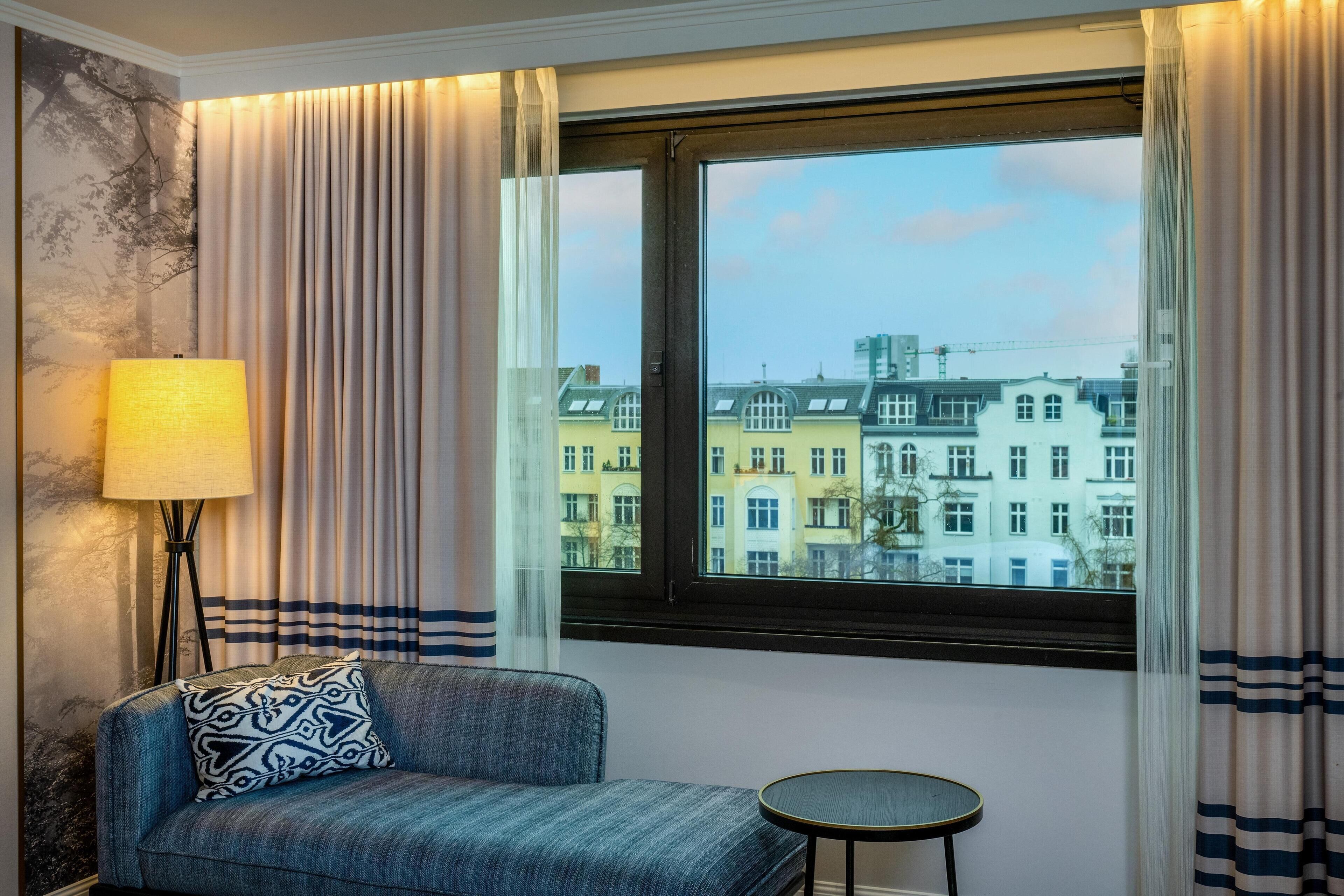 Een kamer met een bank, een klein rond tafeltje, een lamp en een raam met gordijnen.