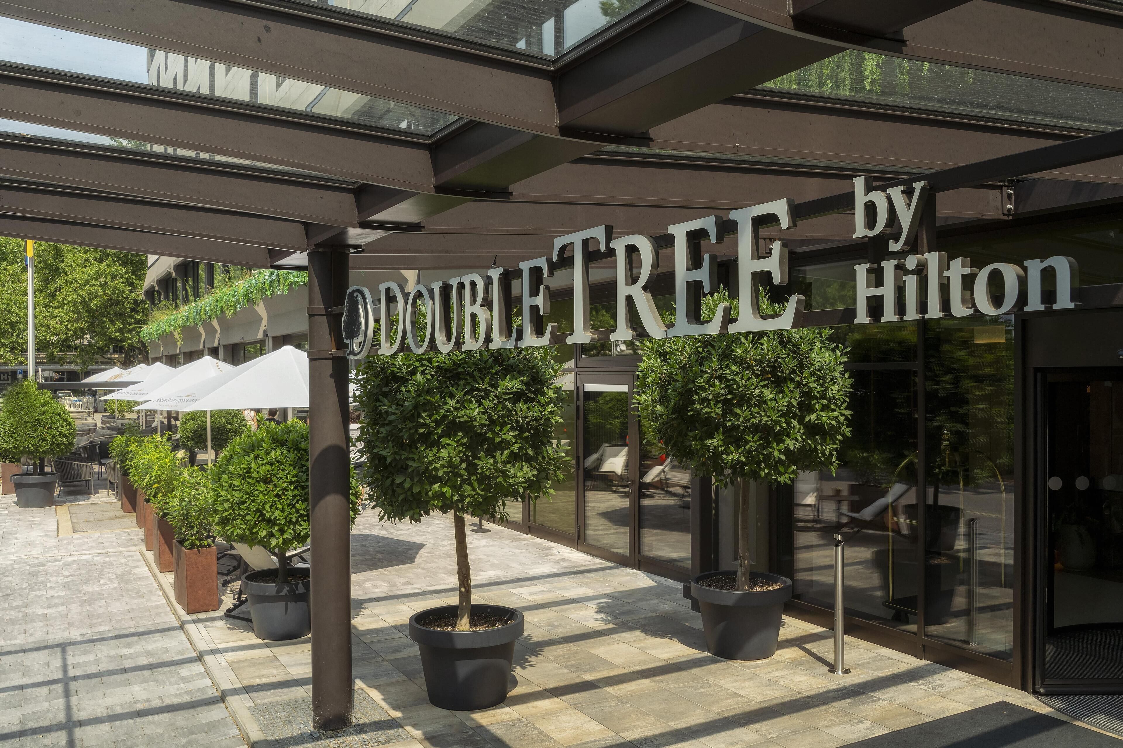 A hotel entrance with "DOUBLETREE by Hilton" signage, potted trees, and outdoor seating.