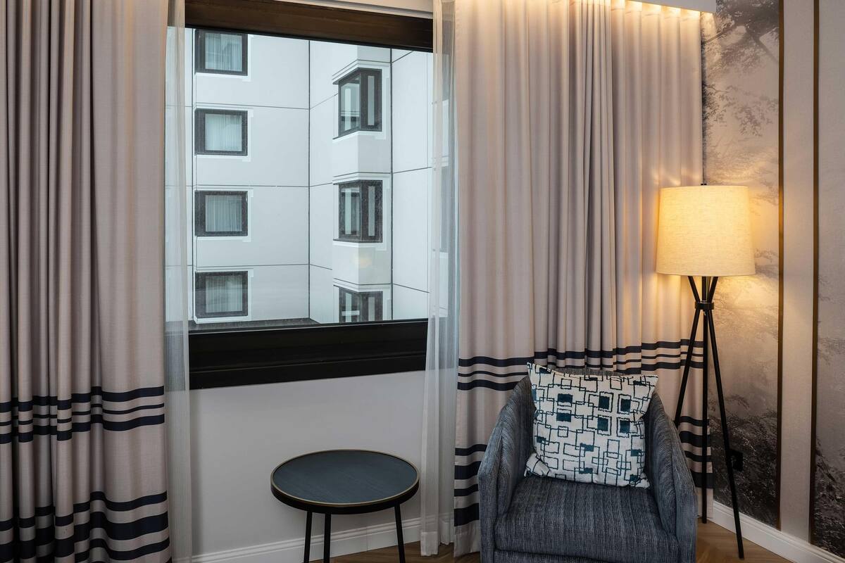 Habitación de hotel con vista a un edificio a través de una ventana, una lámpara de pie, una silla con cojín estampado y una pequeña mesa redonda.