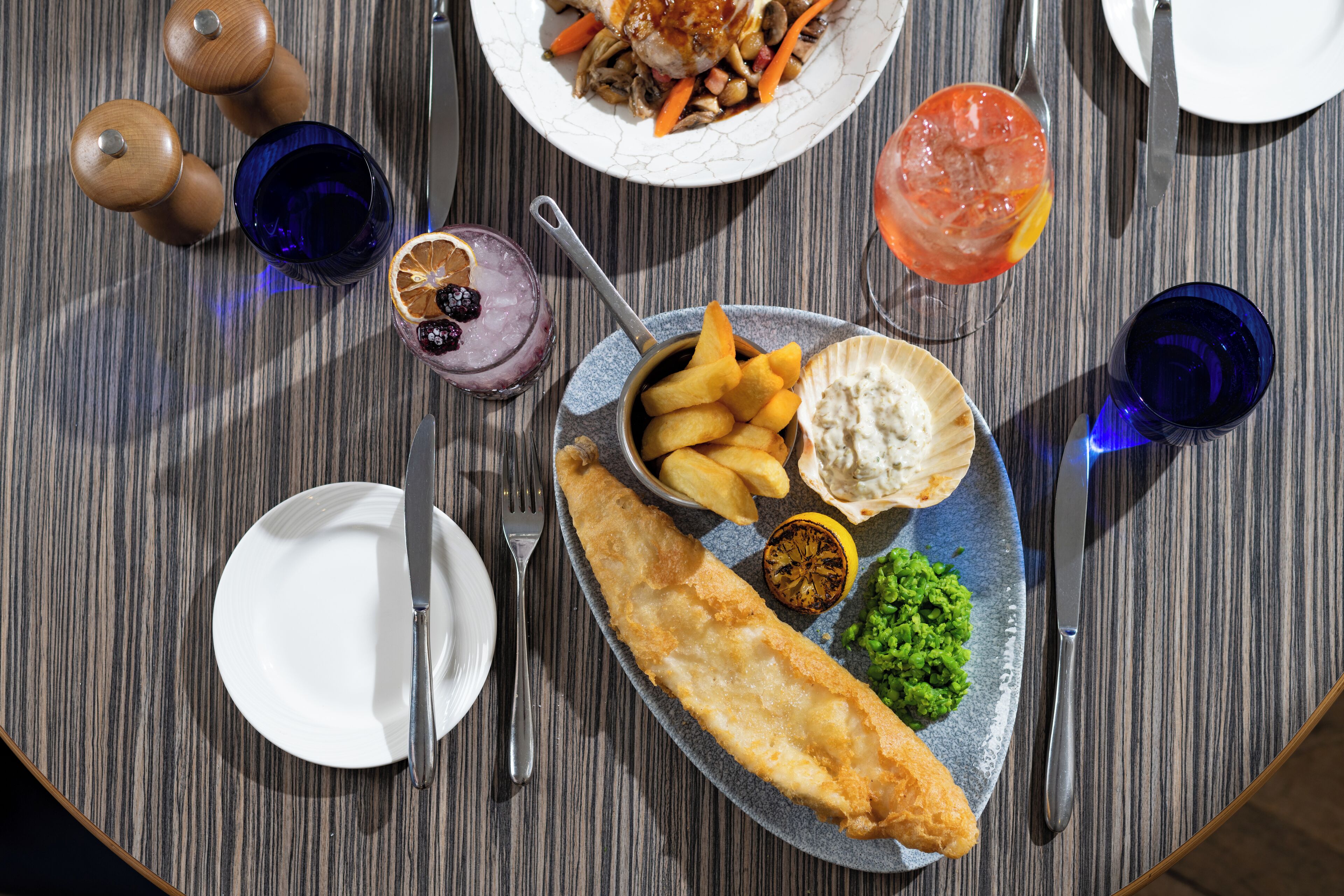A table set with a fish and chips meal, including a side of mushy peas and chips, a glass of a red drink, and a small bowl of pickles.