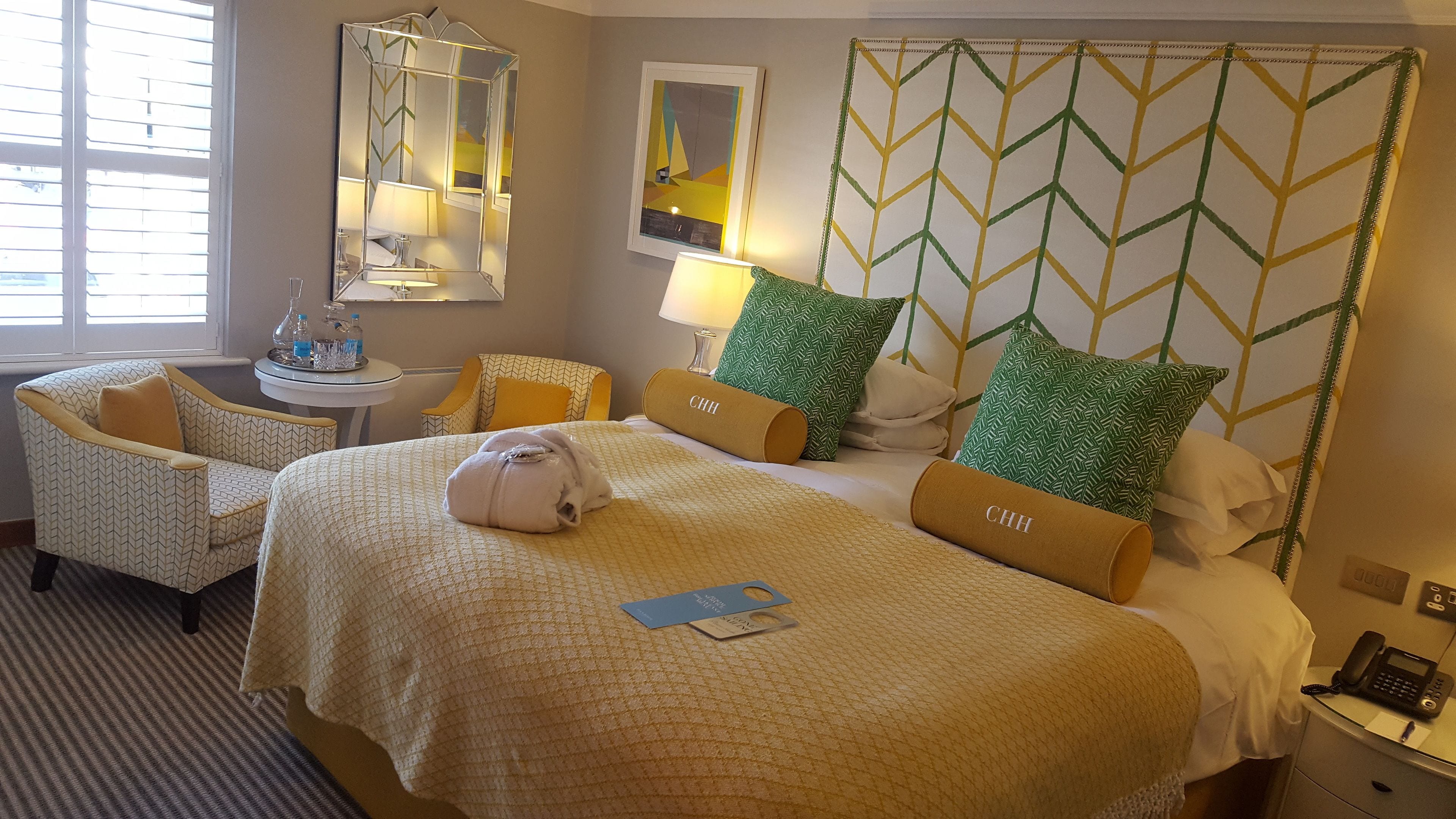 A hotel room with a large bed, a sitting area with a chair and a small table, a mirror, and a wall with a geometric pattern.