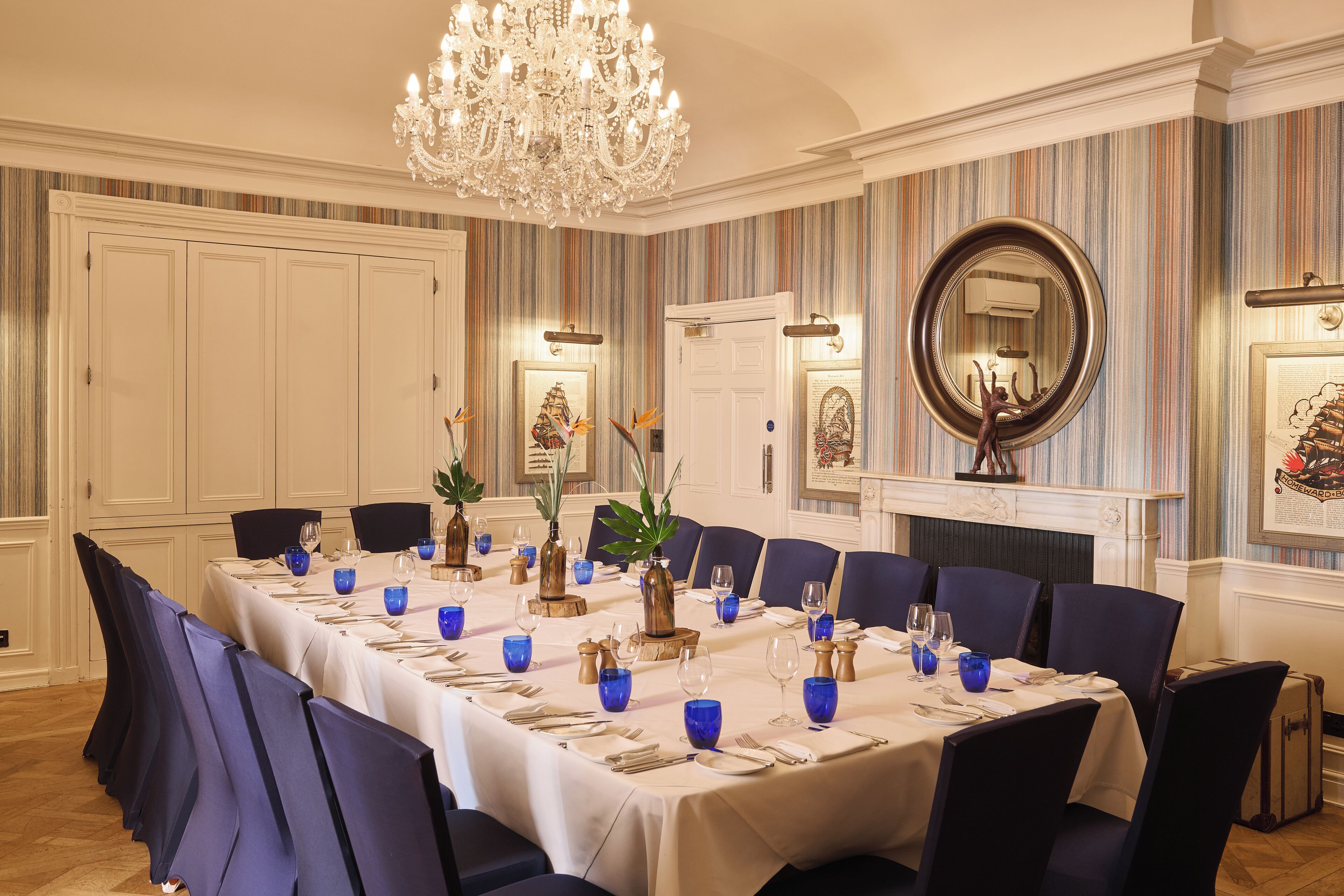 Une salle à manger élégante, avec une longue table dressée pour un repas, entourée de chaises, d’un lustre et d’un papier peint à rayures.