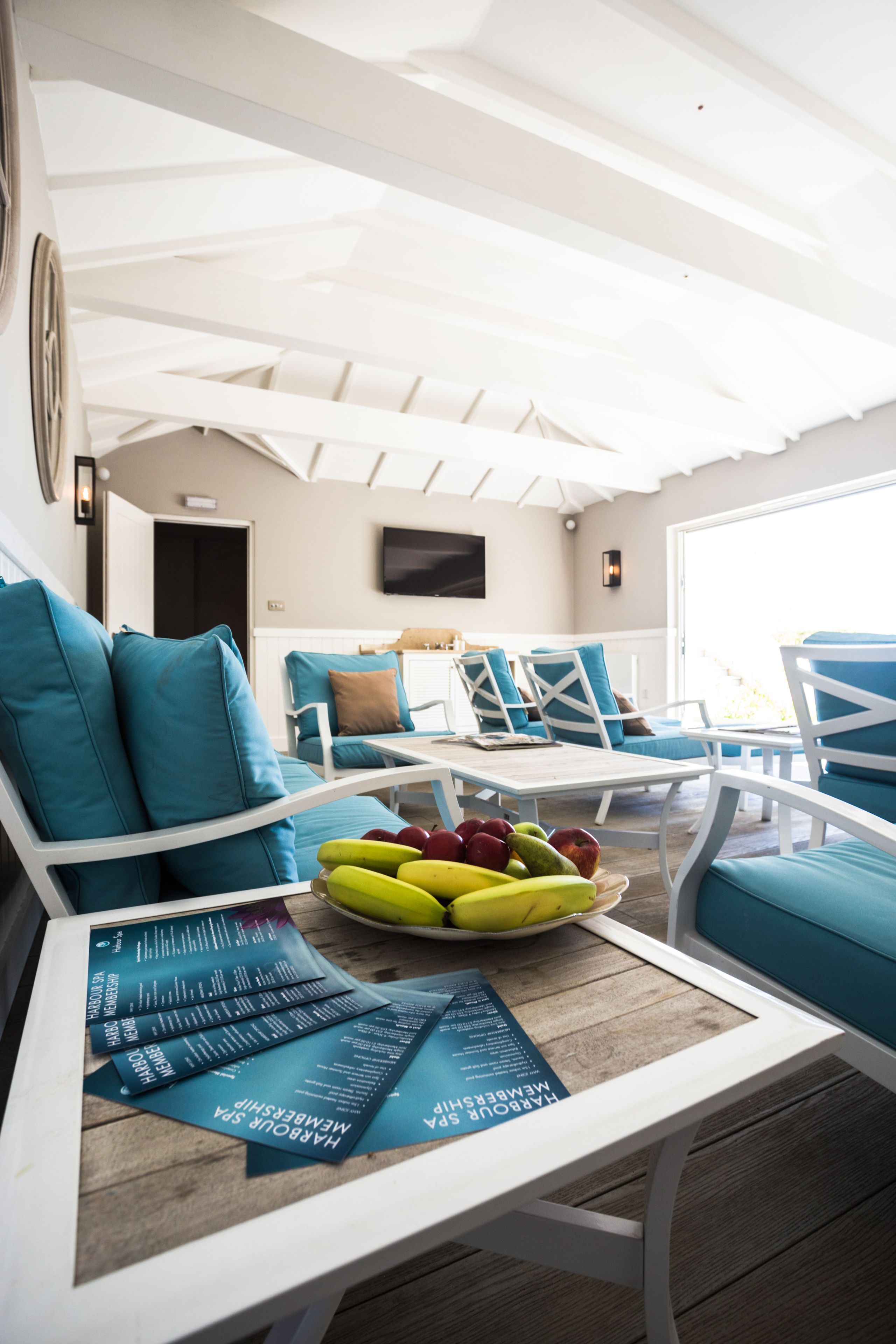 Un'area salotto all'aperto illuminata dal sole, con mobili bianchi e azzurri, un tavolo con una ciotola di frutta e un menù.