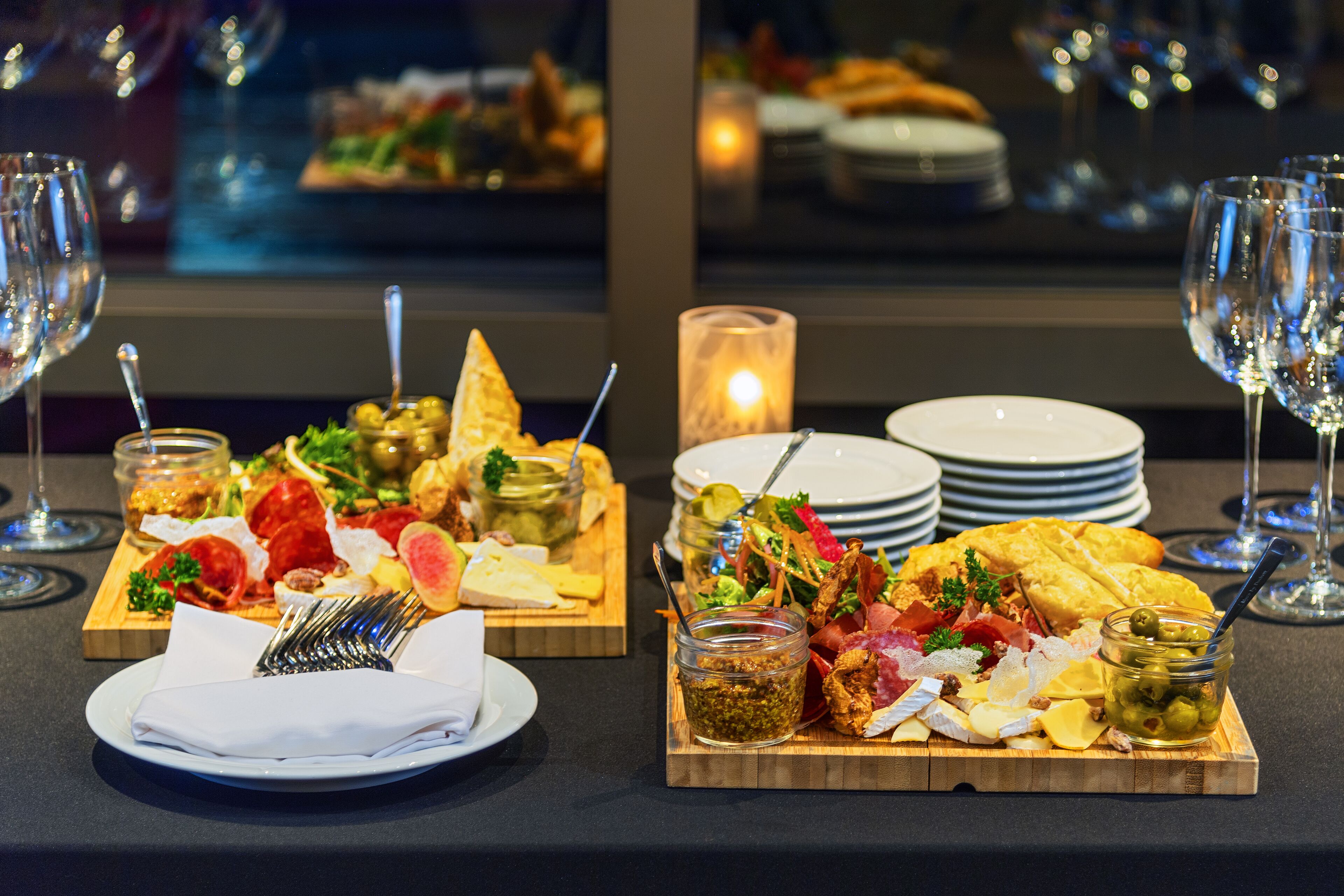 Een eettafel met diverse schalen eten, waaronder kaas, vleeswaren en olijven, en een kaarsje op de achtergrond.