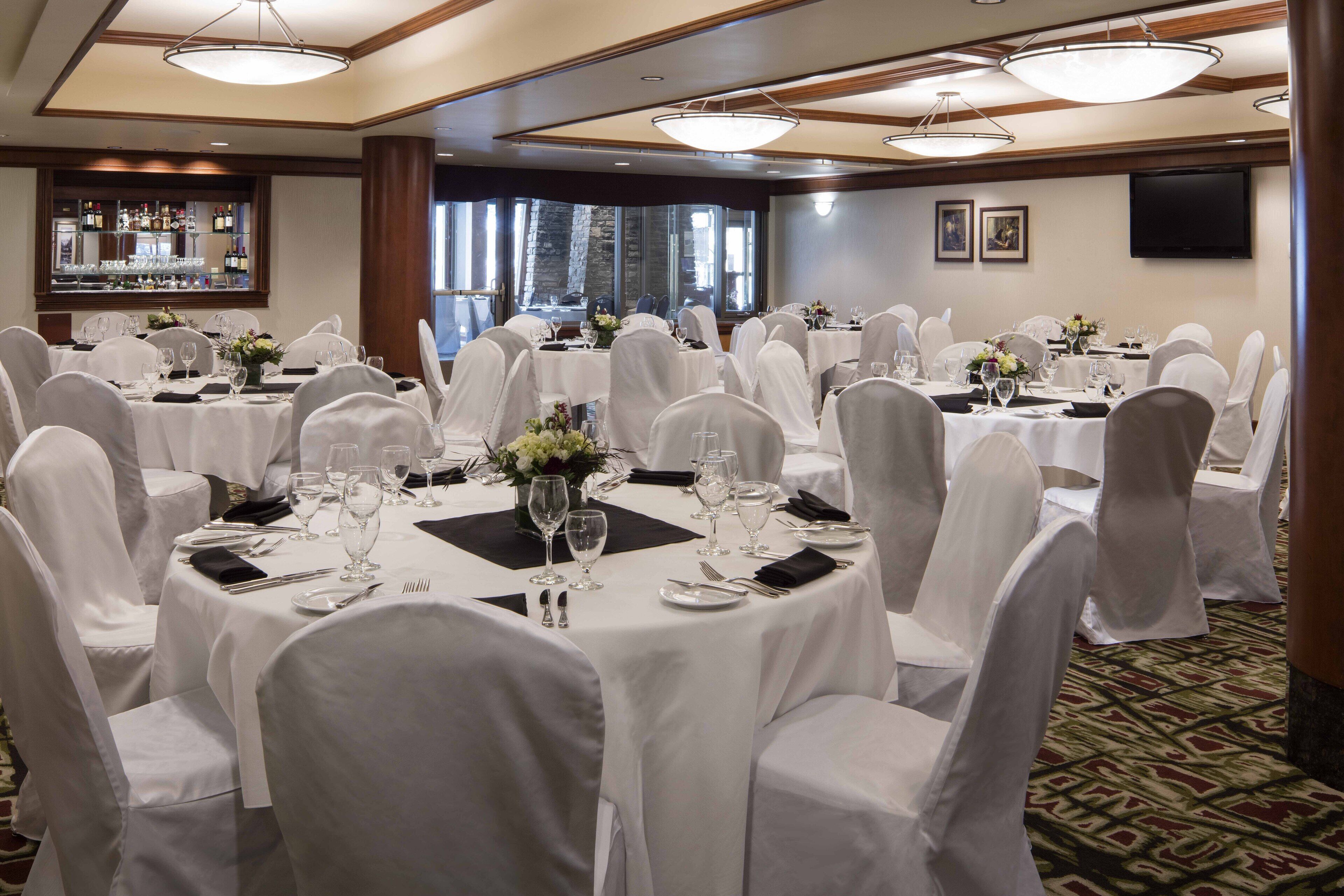 A banquet hall with round tables set for dining, white chairs, and a bar area with bottles.