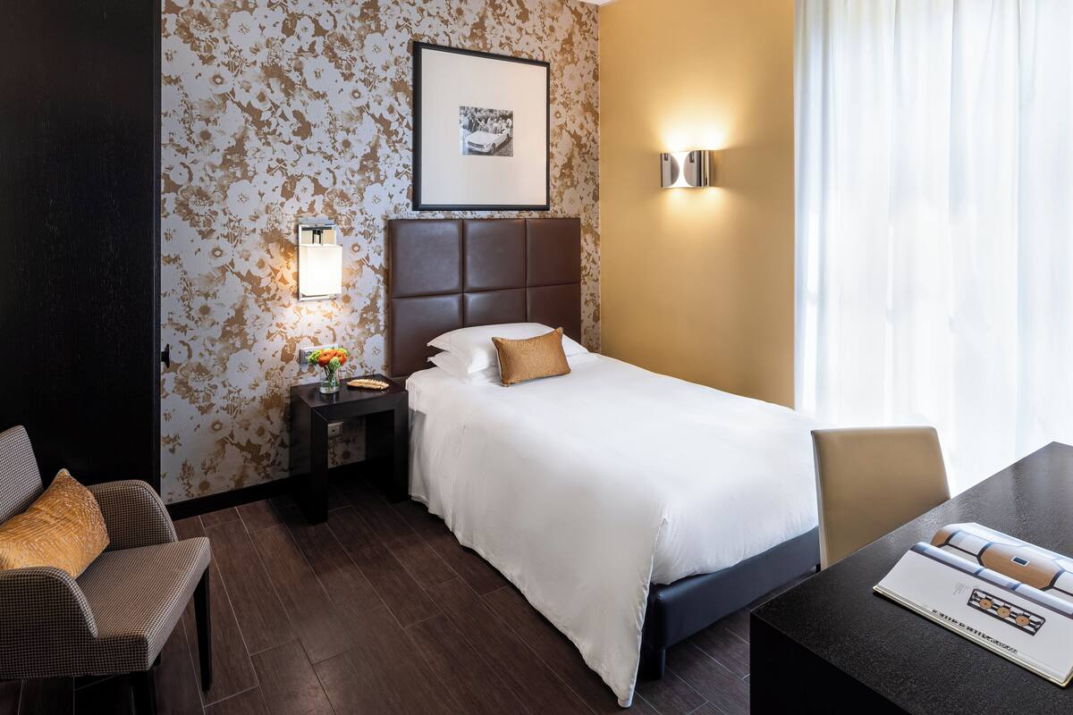 A hotel room with a bed, a nightstand with a lamp, a chair, a table with a magazine, and a wall with a framed picture.