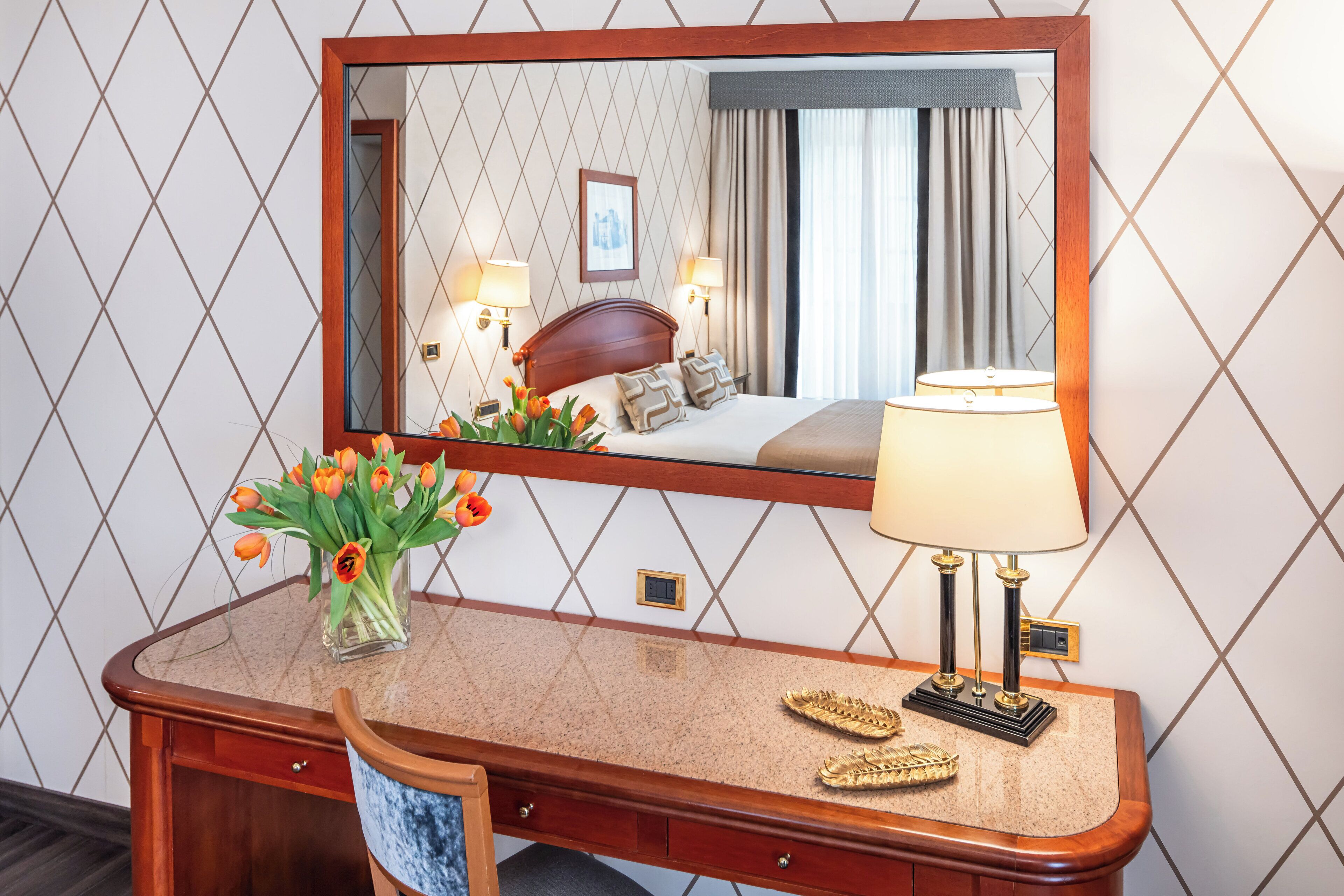 A hotel room with a wooden desk, a mirror, a lamp, and a vase of flowers.