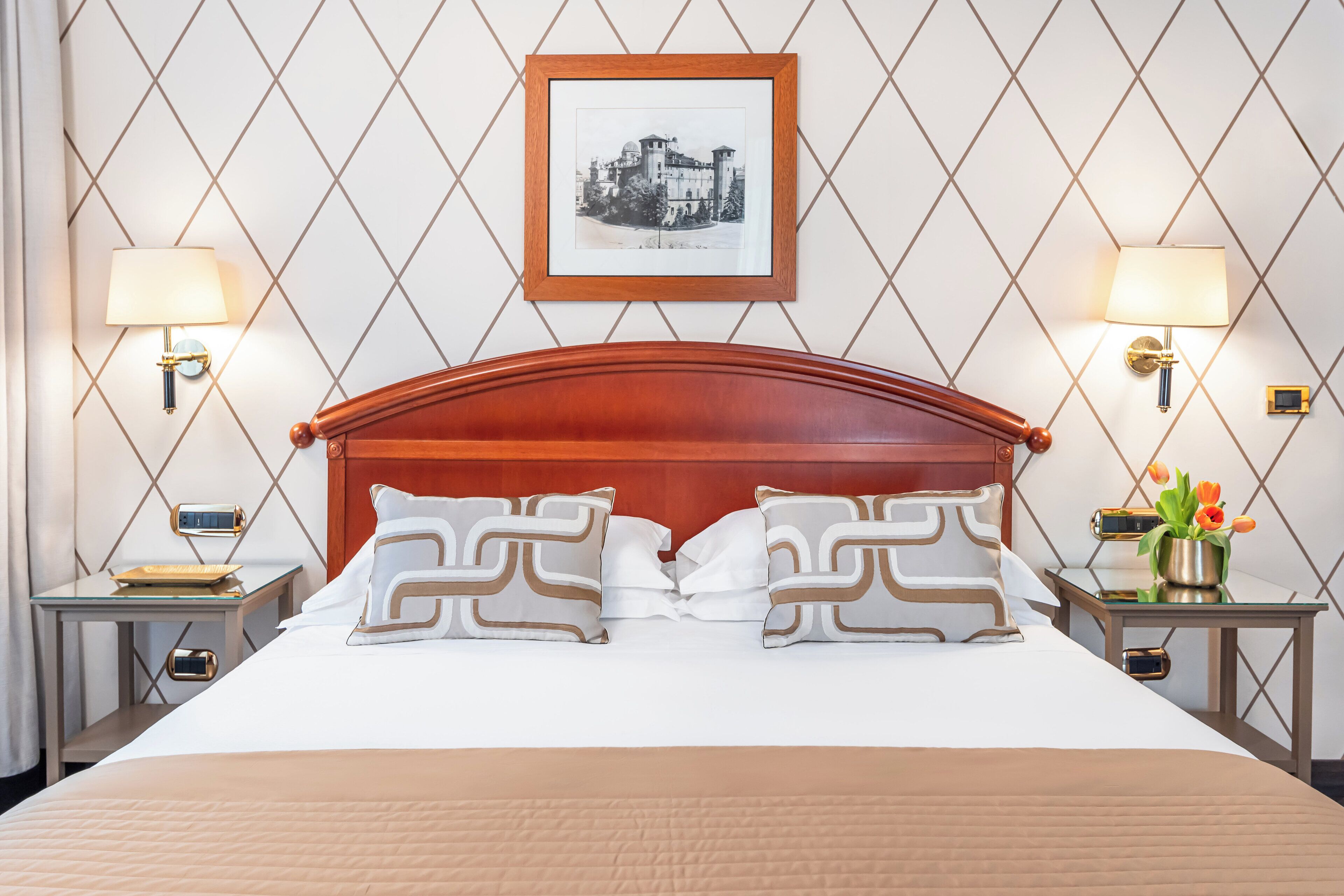 Una habitación de hotel con una cama grande, dos mesitas de noche, un cuadro enmarcado en la pared y una mesita con una lámpara.