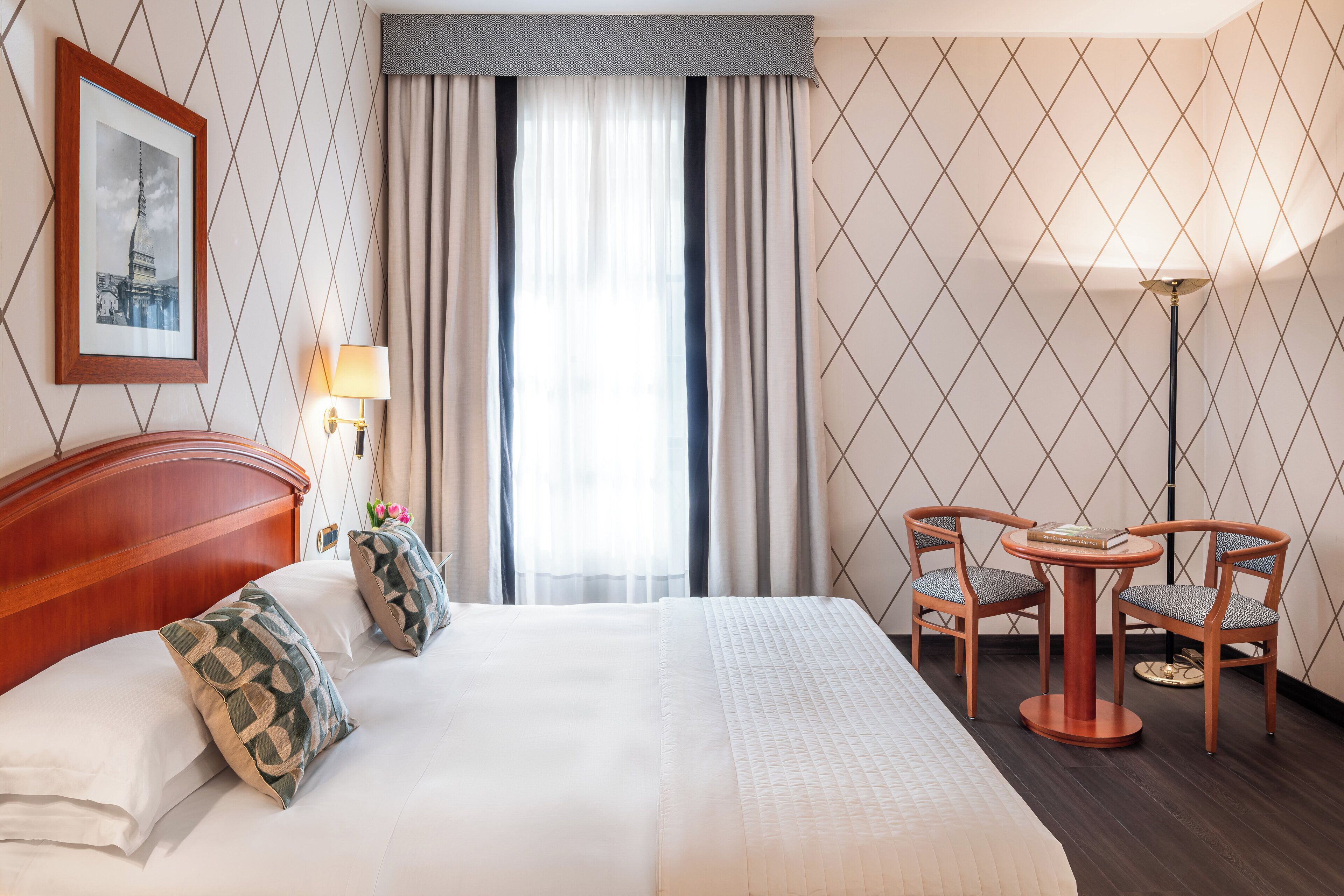 Una camera d'albergo con un letto grande, due sedie attorno a un tavolino, una lampada e un quadro appeso al muro.