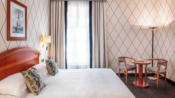 Una camera d'albergo con un letto grande, due sedie attorno a un tavolino, una lampada e un quadro appeso al muro.