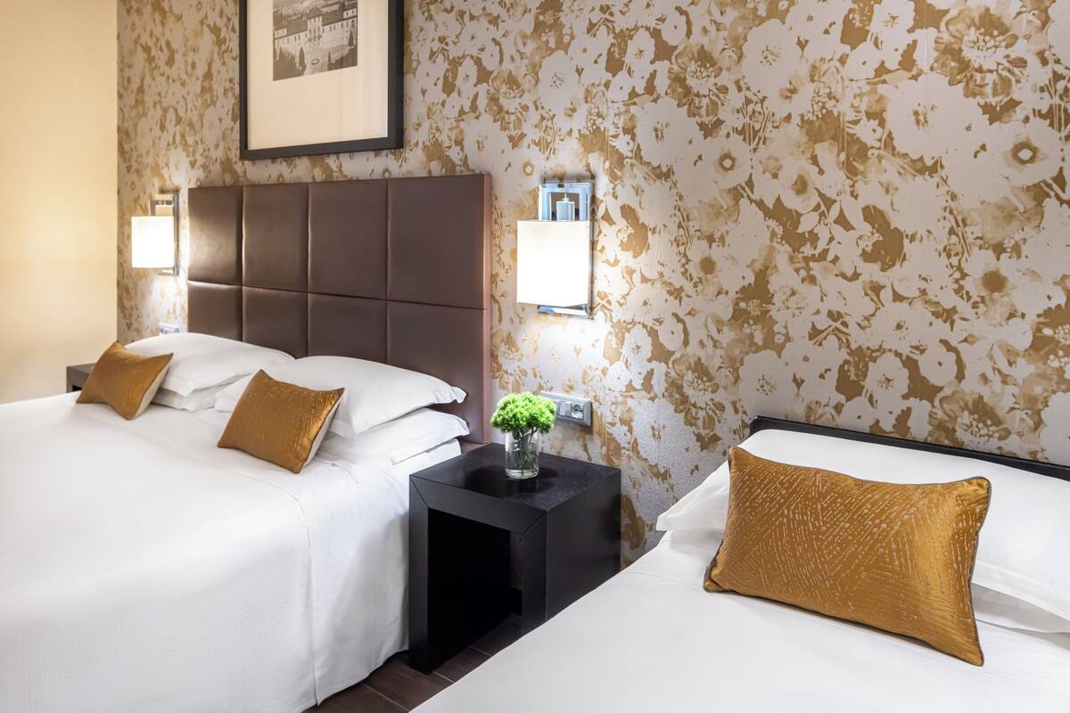 Een hotelkamer met twee bedden, een donker hoofdeinde, een nachtkastje met een bloevas en een wandlamp.