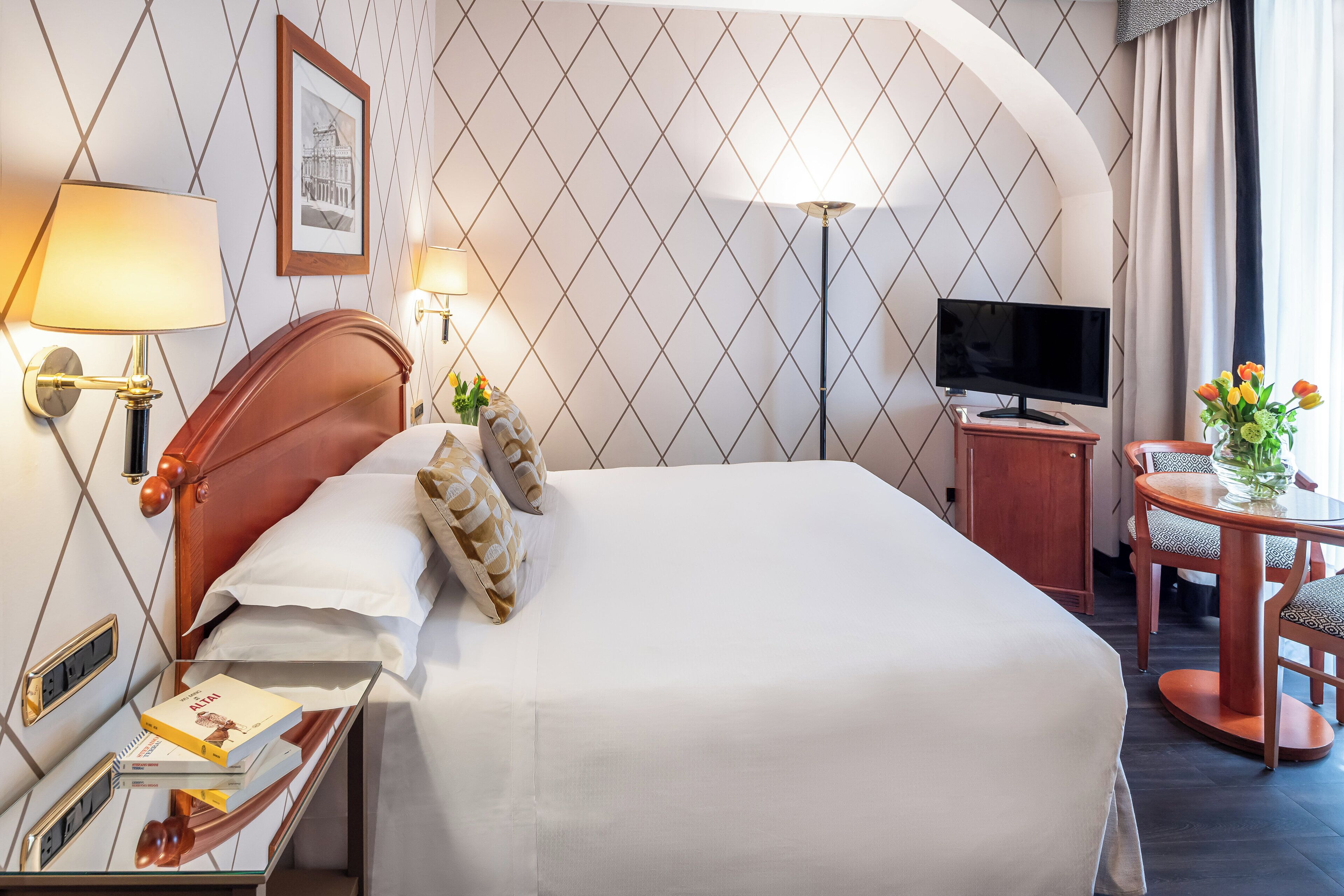 A hotel room with a neatly made bed, a bedside table with a lamp, a TV, and a small round table with chairs.