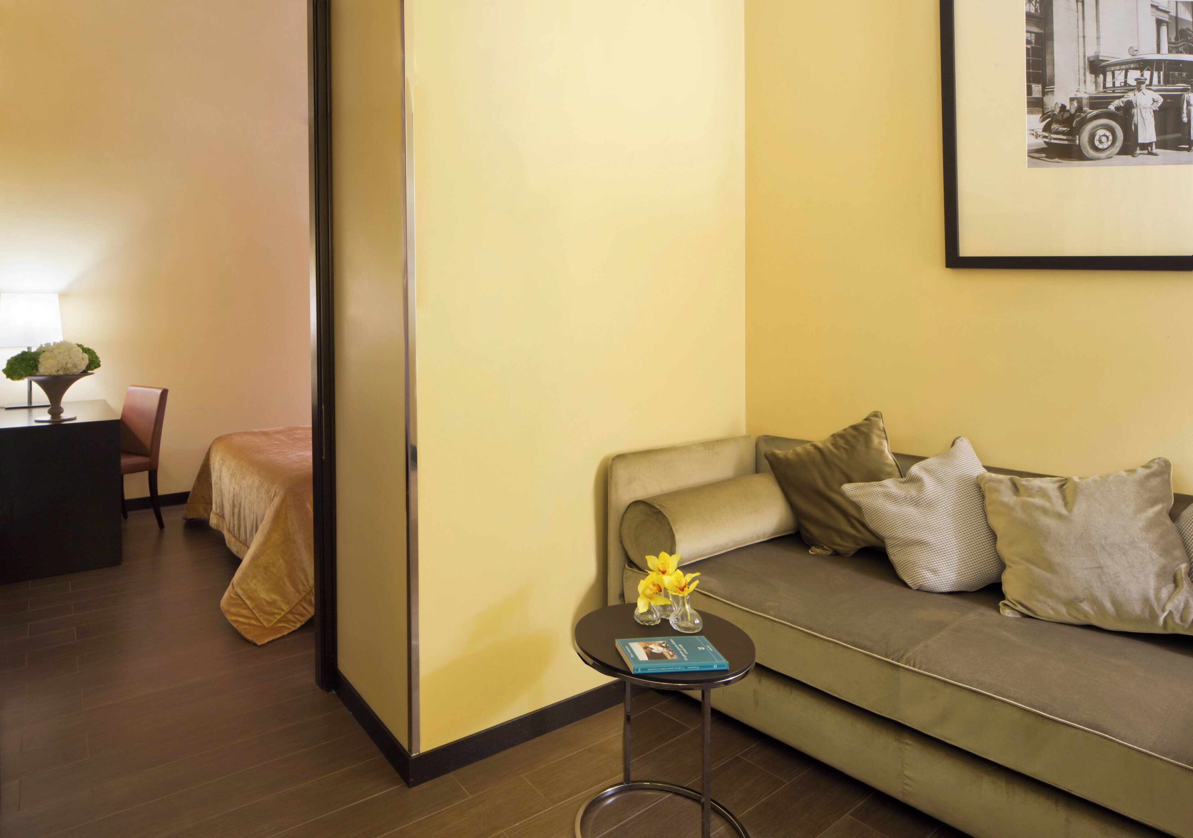 A hotel room with a sofa, a small table with a vase of flowers, a framed picture on the wall, and a bed with a headboard.