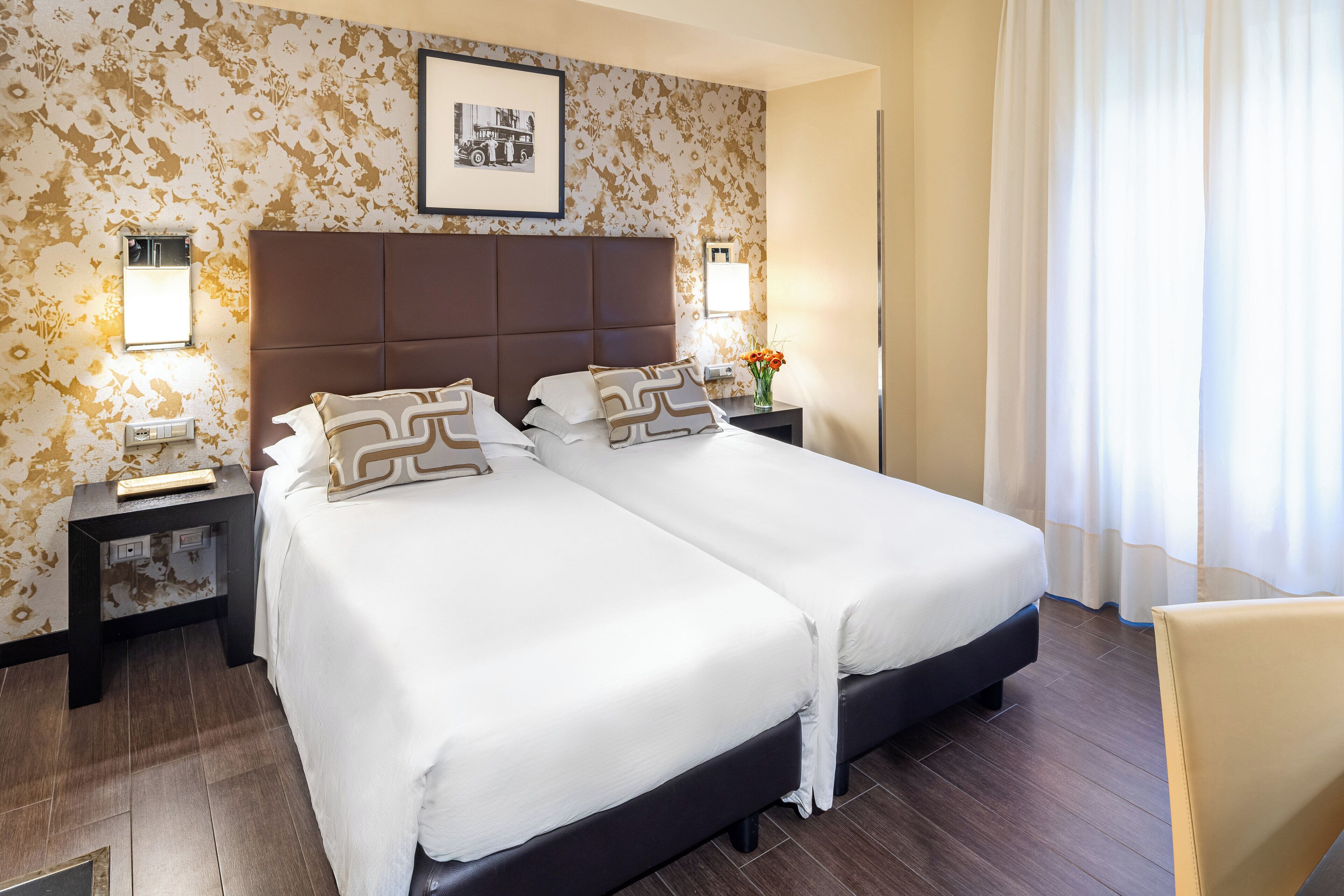 Habitació d'hotel amb dos llits, un capçal, taules de nit, una cadira i un quadre a la paret.