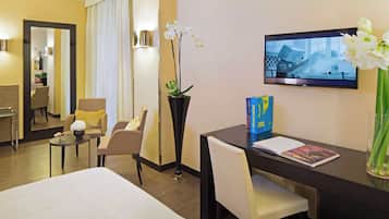Una camera d'albergo con un letto, una scrivania con una sedia, una televisione e un vaso con fiori.