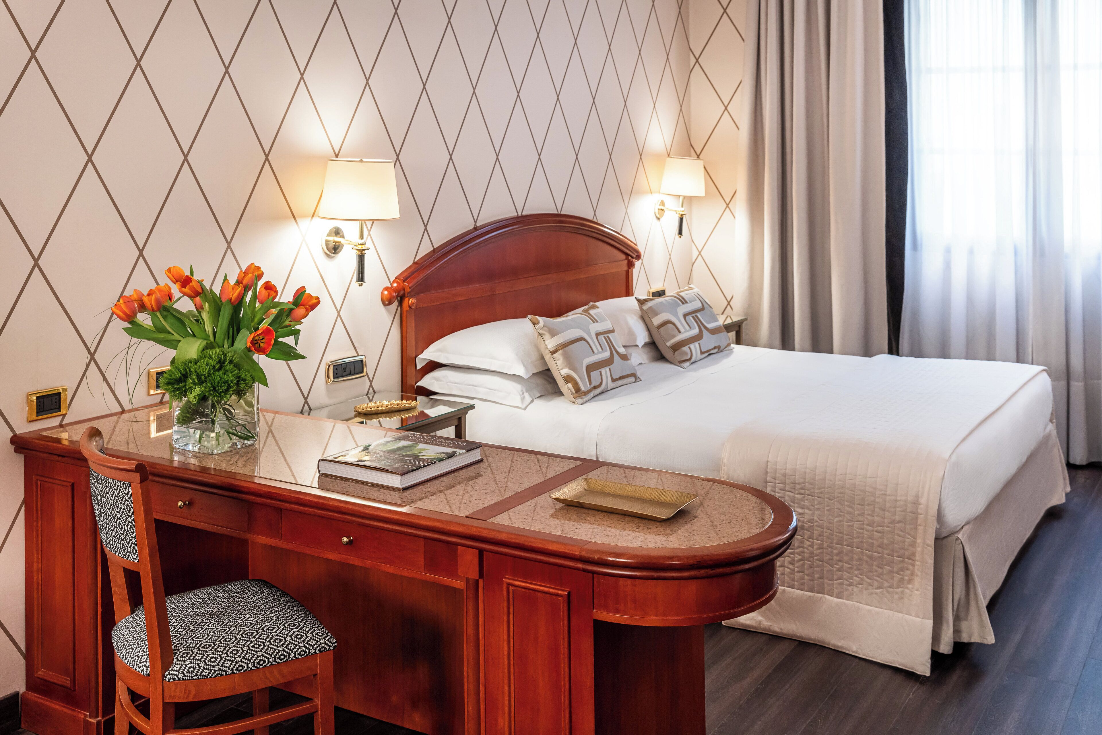 A hotel room with a wooden desk, a chair, a bed with white bedding, and two wall-mounted lamps.