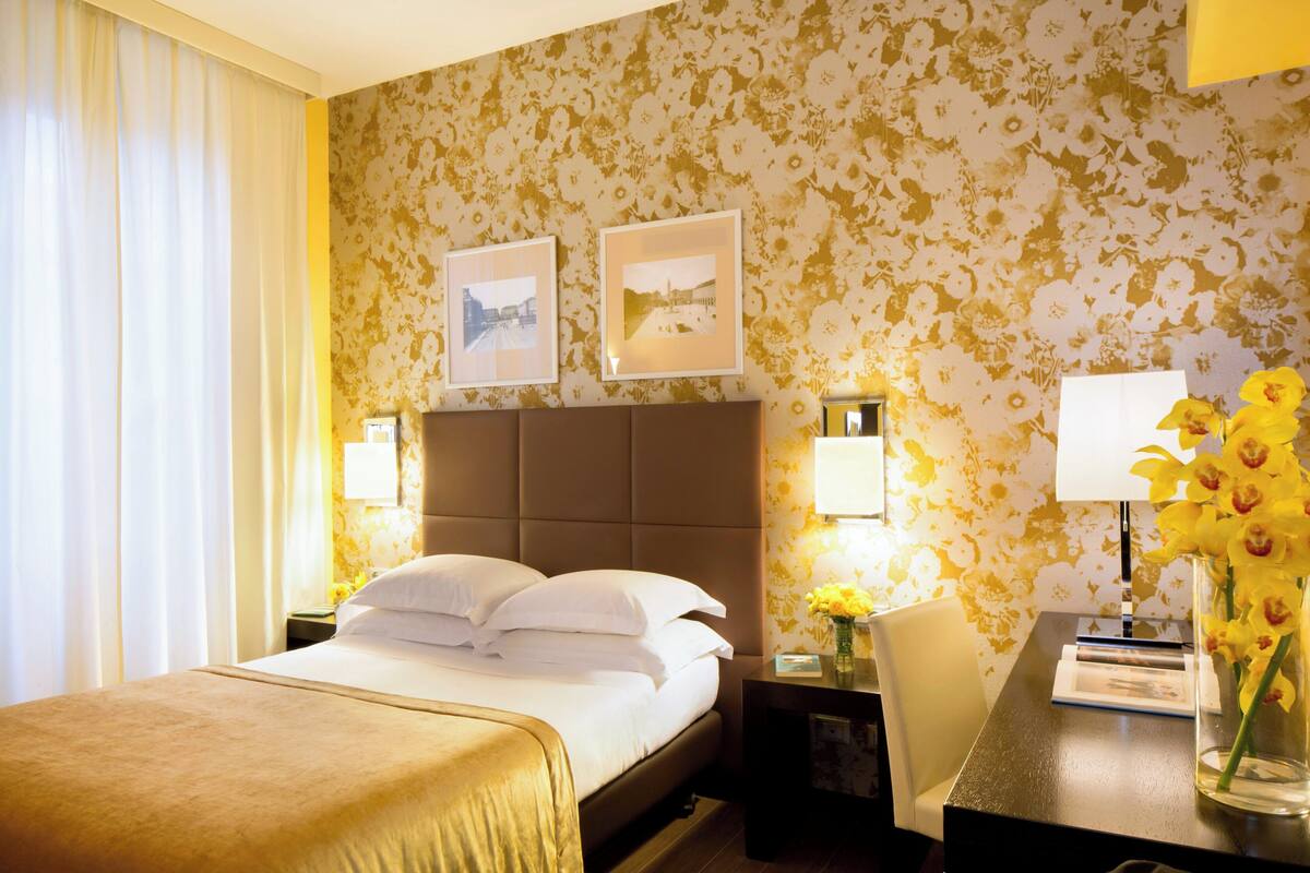 Een hotelkamer met een groot bed, een nachtkastje met een lamp, een bureau met een bloevas en twee ingelijste foto's aan de muur.