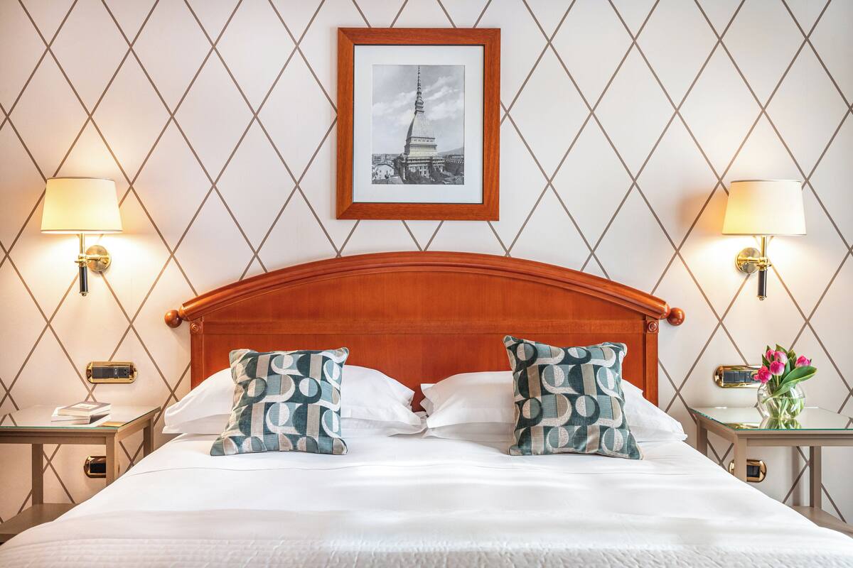 Hotelkamer met een houten hoofdbord, een ingelijst kunstwerk aan de muur, twee nachtkastjes met lampen en gestreepte kussens.