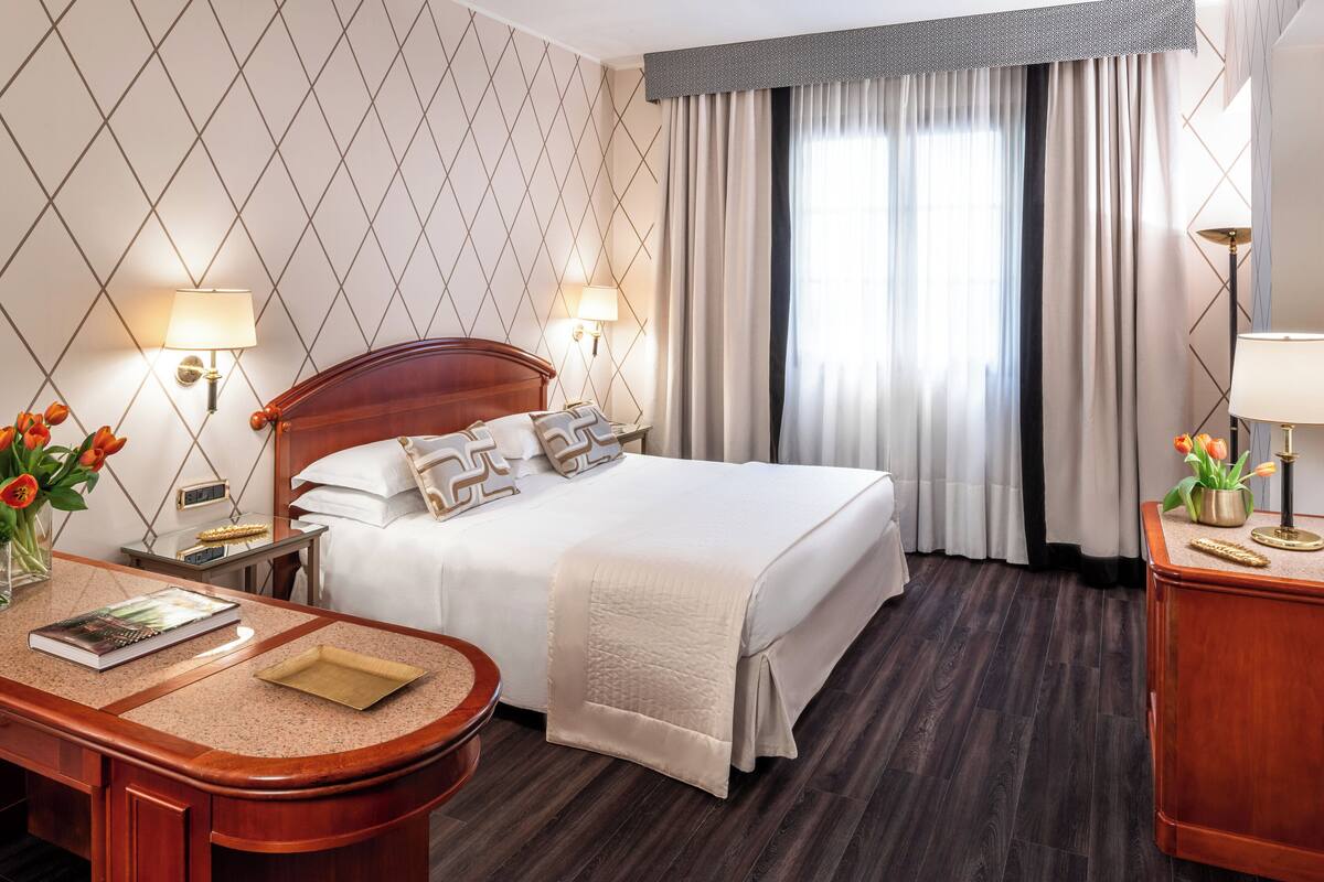 A hotel room with a large bed, two bedside lamps, a desk with a chair, a small table with a vase of tulips, and a window with curtains.