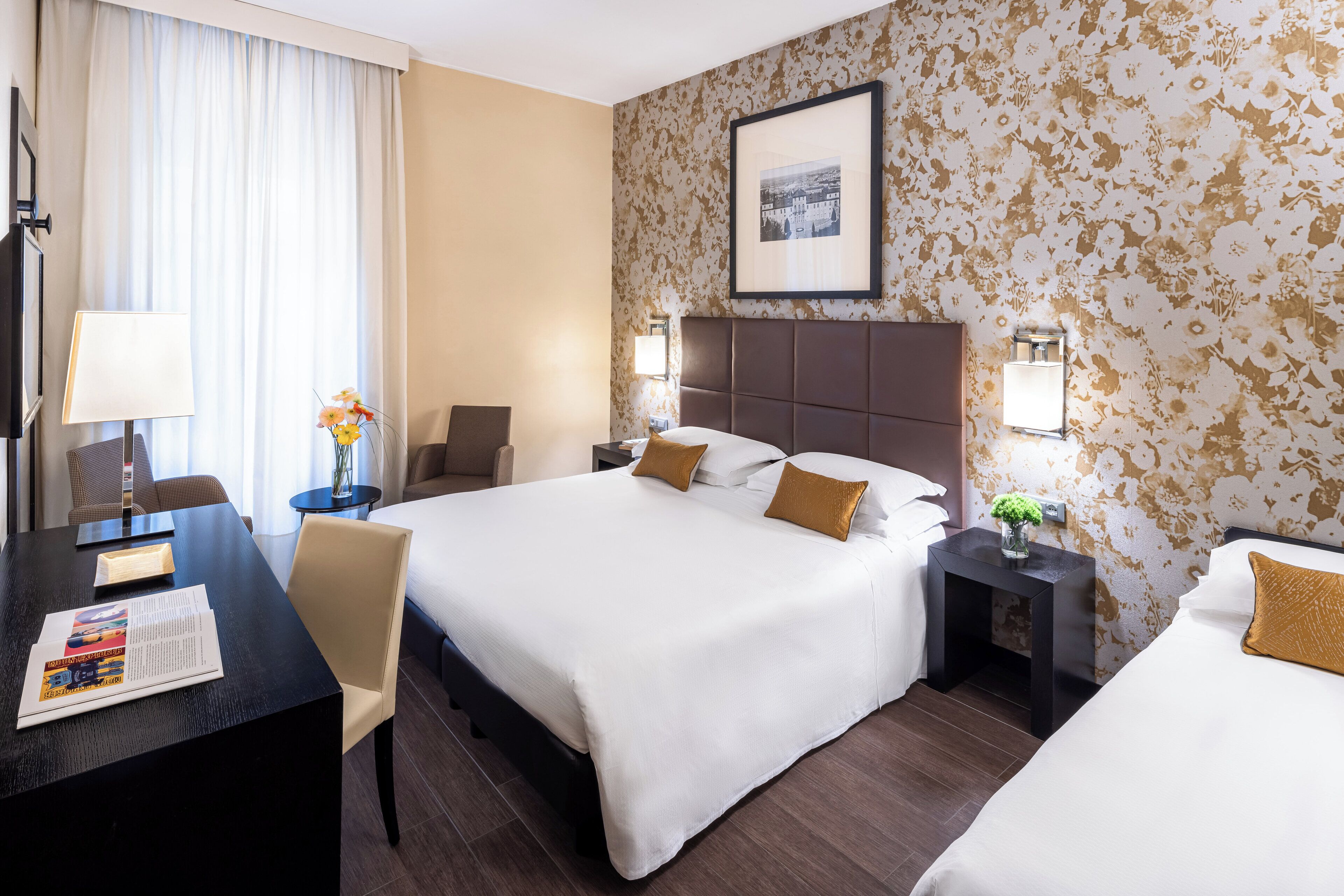 Una camera d'albergo con due letti, una scrivania, una sedia e un comodino con una lampada.